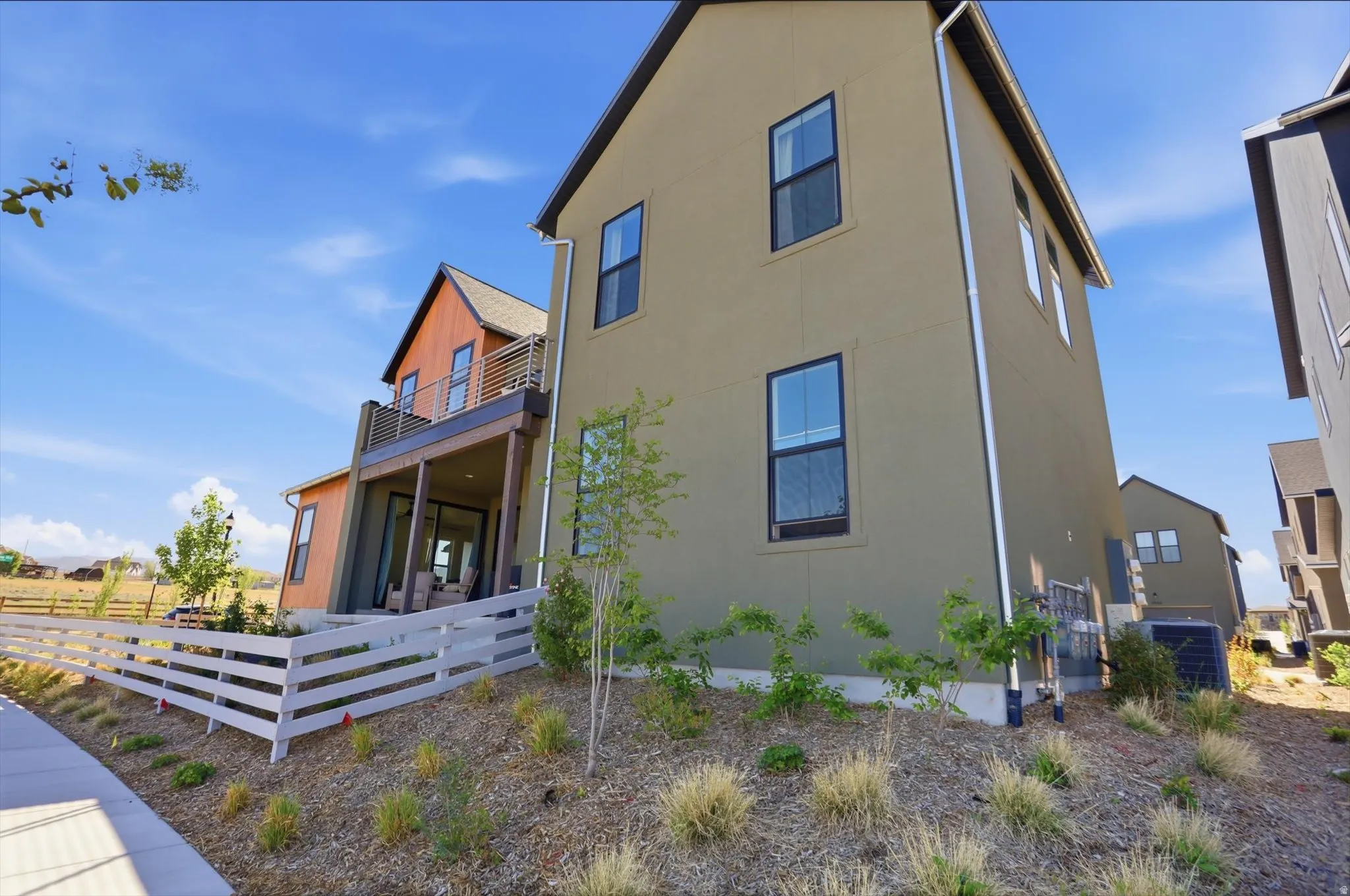 Modern exterior featuring a covered patio, upper-level balcony with metal railings, and a white picket fence