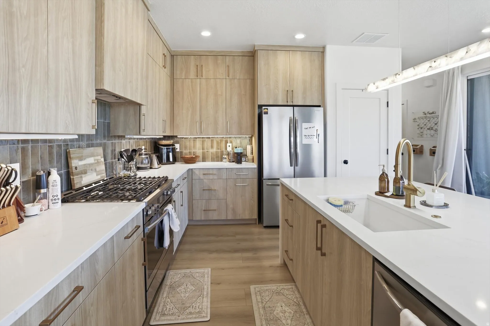 Modern kitchen featuring wood-finish cabinetry, white countertops, stainless steel appliances, a gas range, and a single basin sink with a gold-tone faucet