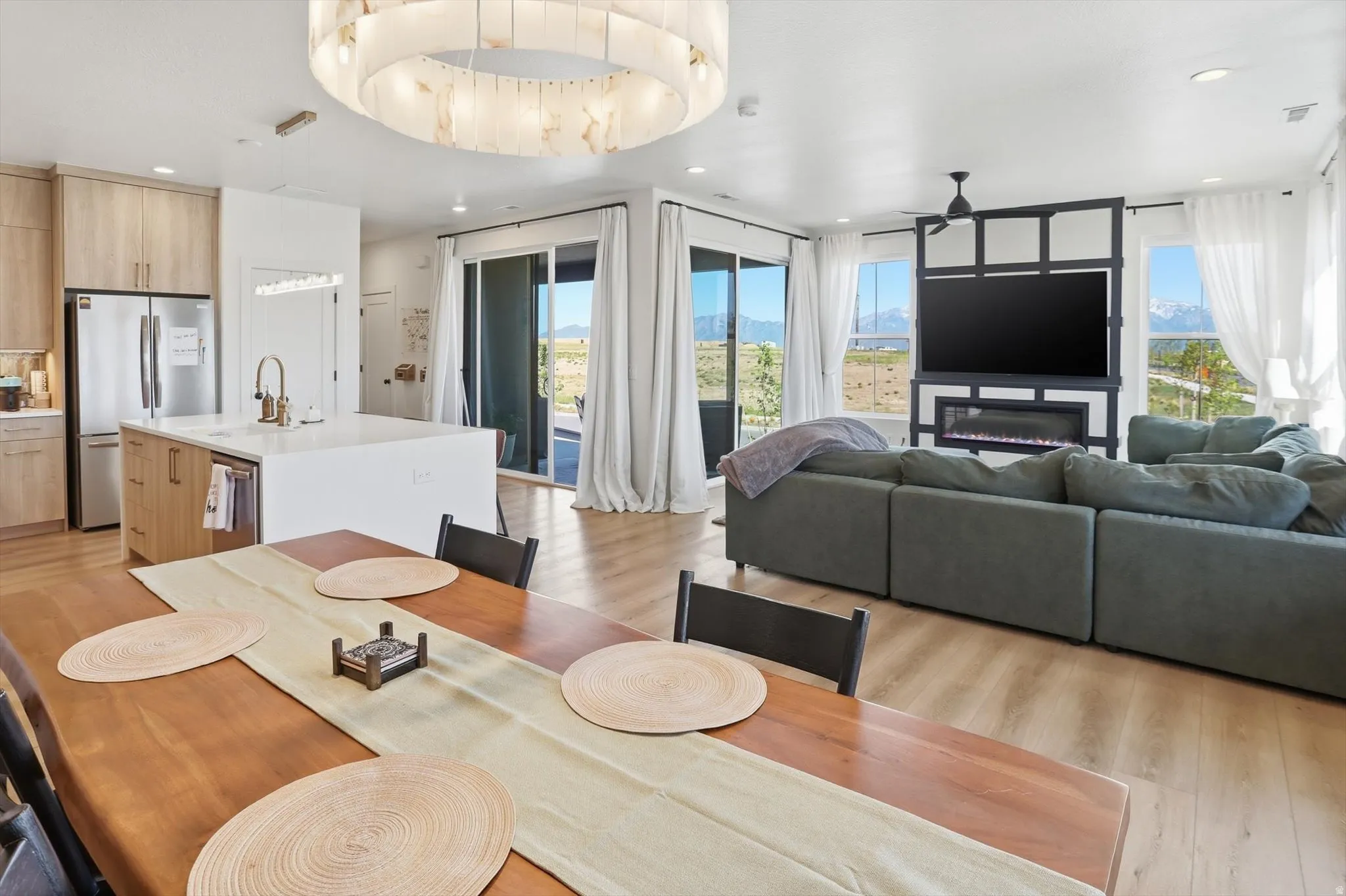 Open-concept living area featuring light wood-finish flooring, a built-in electric fireplace with media center, and expansive windows showcasing mountain scenery