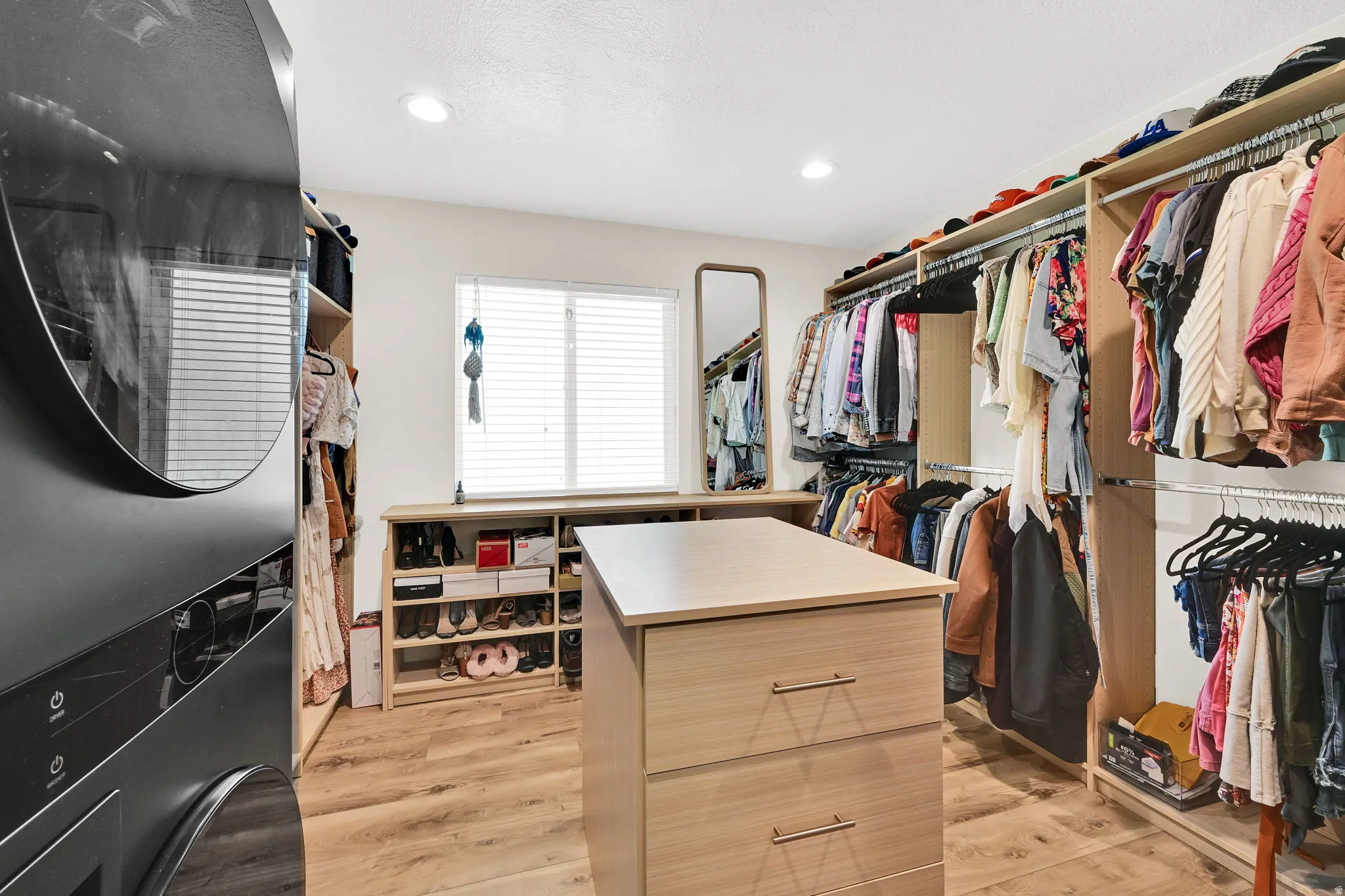 Custom primary suite closet system featuring a central island with integrated drawers, electrical charging station, wood-finish flooring, a stacked washer and dryer unit, and LG Styler steaming closet.