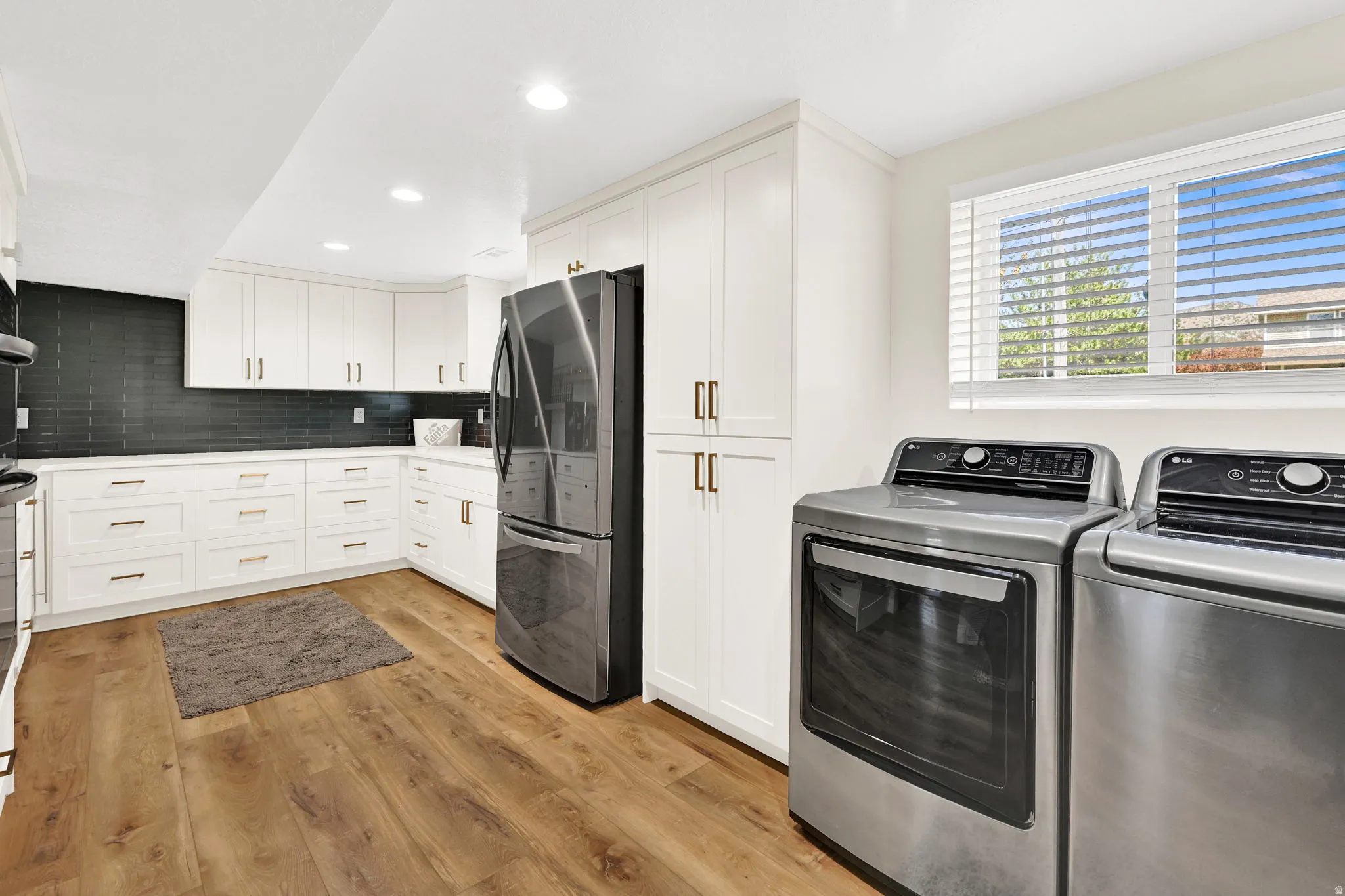 Hidden butler pantry with laundry area featuring wood-finish flooring, white cabinetry with gold-tone hardware, dark subway tile backsplash, and stainless steel appliances