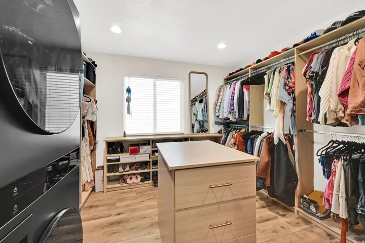 Custom primary suite closet system featuring a central island with integrated drawers, electrical charging station, wood-finish flooring, a stacked washer and dryer unit, and LG Styler steaming closet.