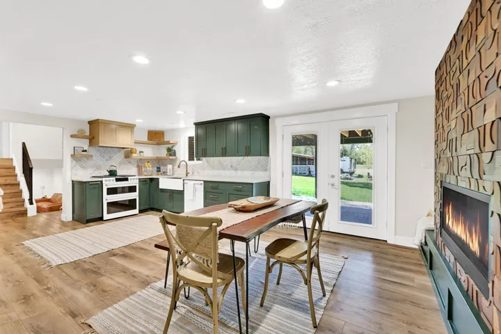 Modern kitchen and dining area featuring deep green cabinetry, white appliances, wood-finish flooring, a herringbone tile backsplash, and a contemporary fireplace with a custom teak wood fireplace.