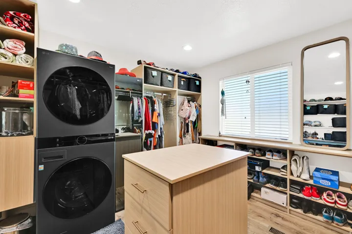 Custom primary suite closet system featuring a central island with integrated drawers, electrical charging station, wood-finish flooring, a stacked washer and dryer unit, and LG Styler steaming closet.
