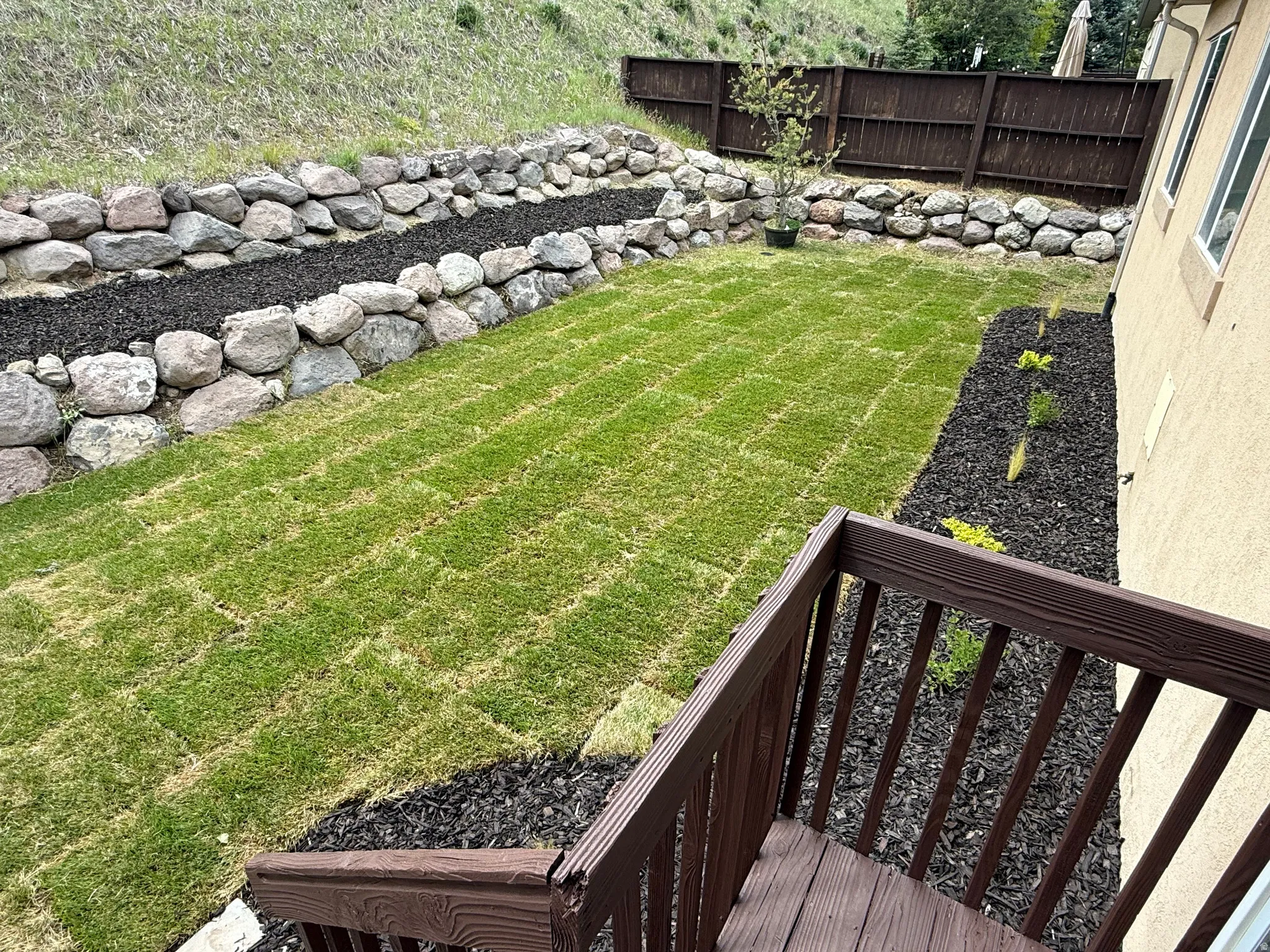 Tiered backyard featuring new sod, dark mulch beds with plantings, and a stone retaining wall