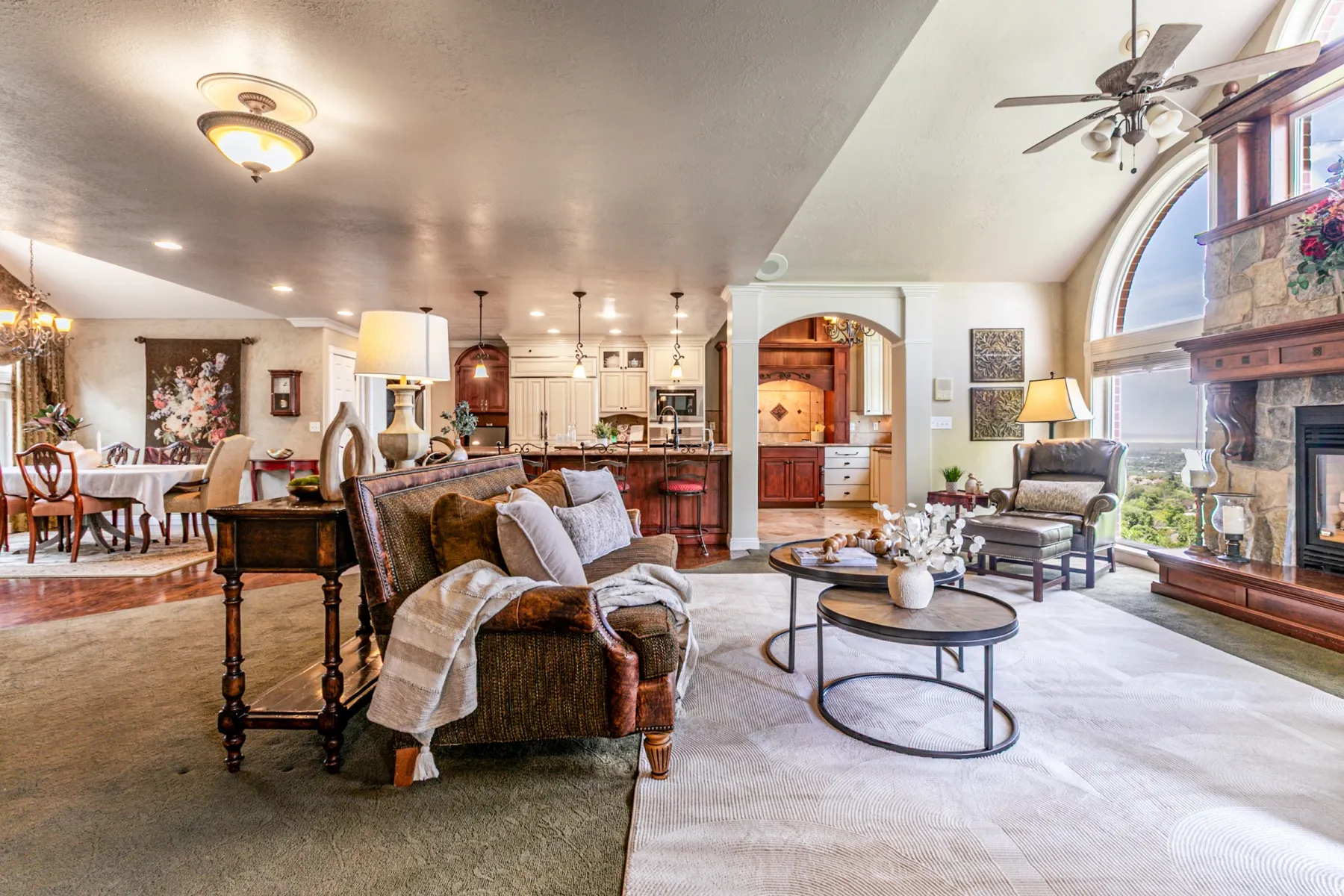 Open-concept living area featuring a stone fireplace with wood mantel, a large arched window, vaulted ceilings, and recessed lighting, dark wood hearth.