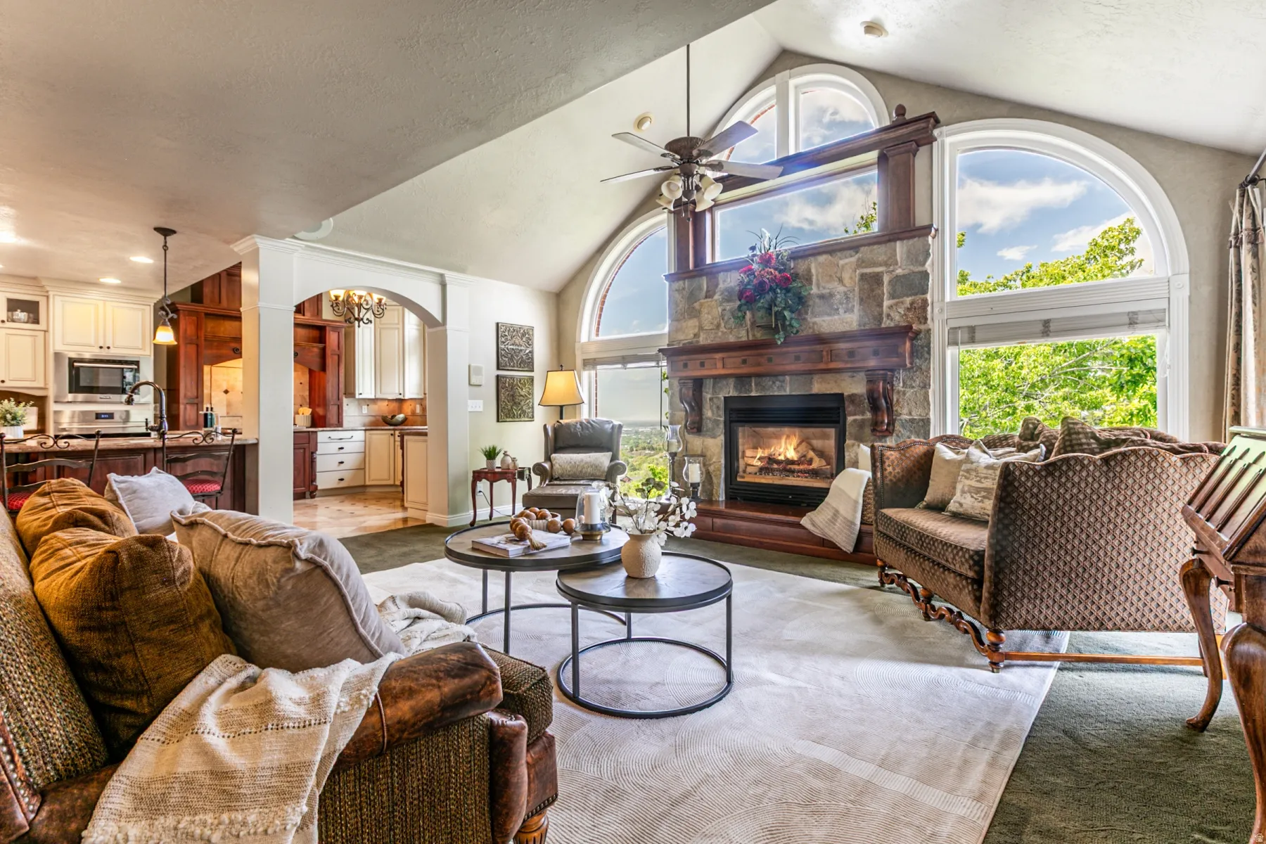 Great room featuring a stone fireplace with a wood mantle, vaulted ceiling with a fan, and expansive arched windows