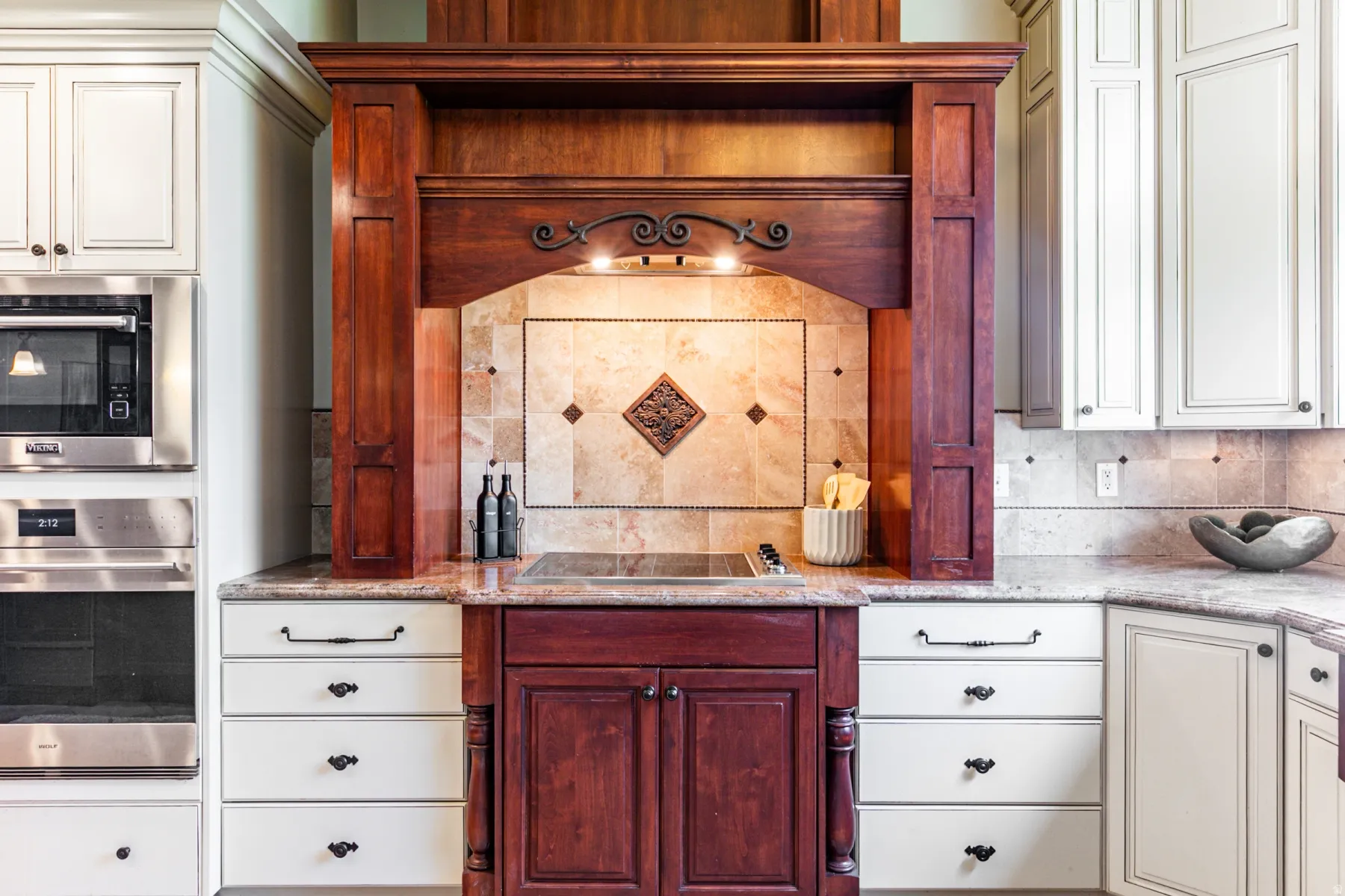 Custom wood range hood with integrated lighting, tile backsplash with decorative accent, and a built-in cooktop