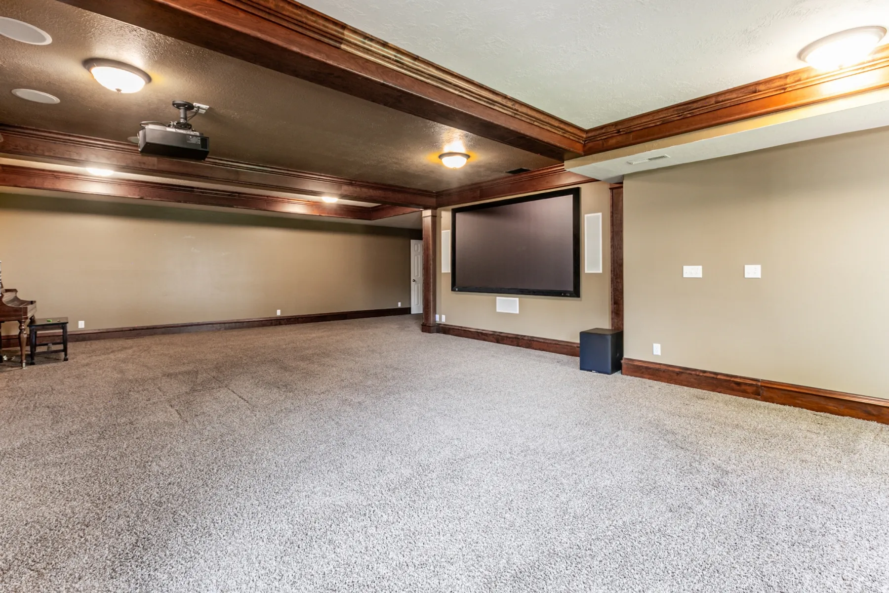 Spacious entertainment room featuring a large projection screen, ceiling-mounted projector, in-ceiling speakers, wood-finish beams, and neutral-toned wall paint