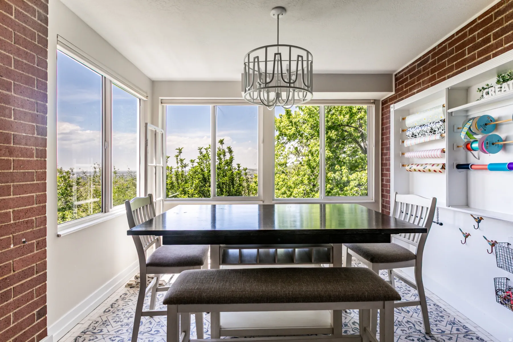 Dining area featuring a dark-toned table, a contemporary chandelier, large windows, exposed brick accents, and patterned floor tiling