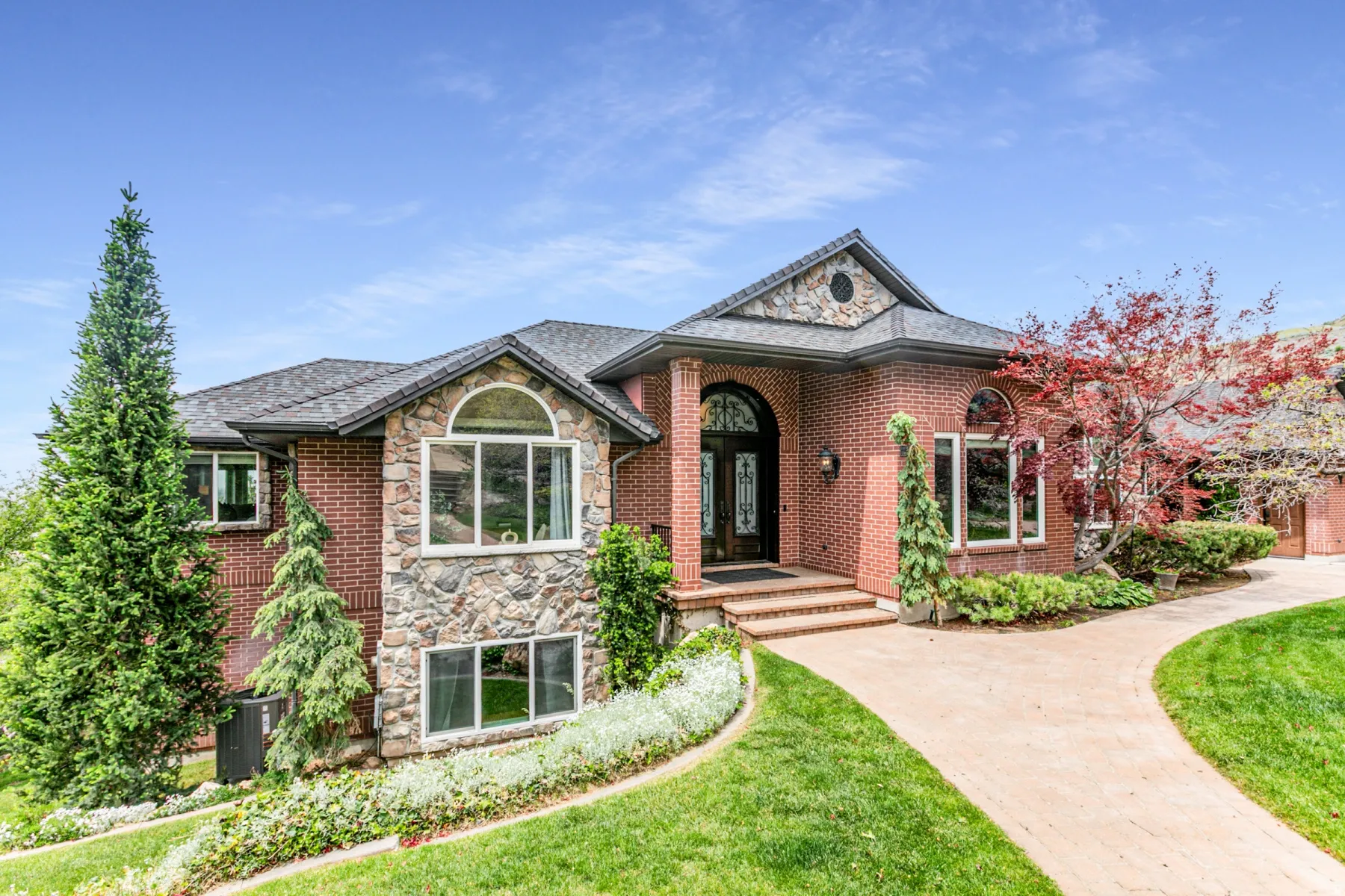 Brick and stone exterior featuring an arched entryway with a decorative glass door, a paved walkway, a shingled roof, and mature landscaping