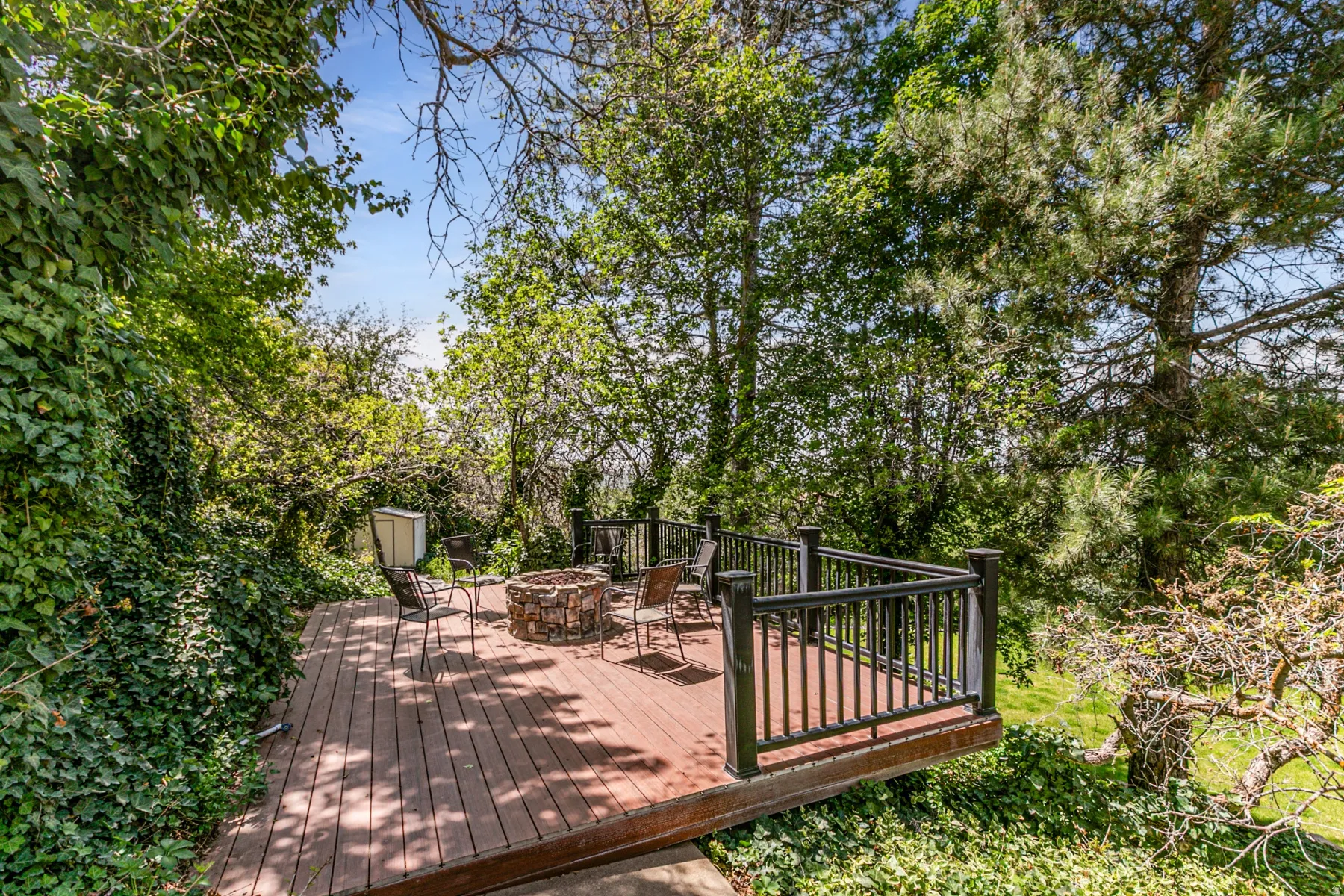Expansive wood-finish deck featuring a stone-clad fire pit, black metal railings, and lush surrounding foliage