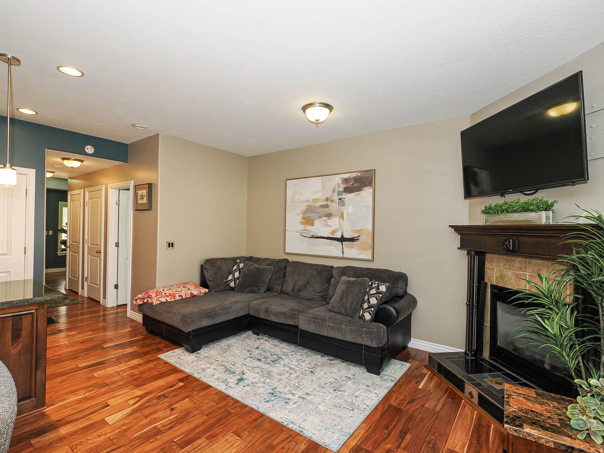 Living area featuring wood-finish flooring, a tiled fireplace with a dark wood mantel, recessed lighting, and a kitchen pass-through with pendant lighting