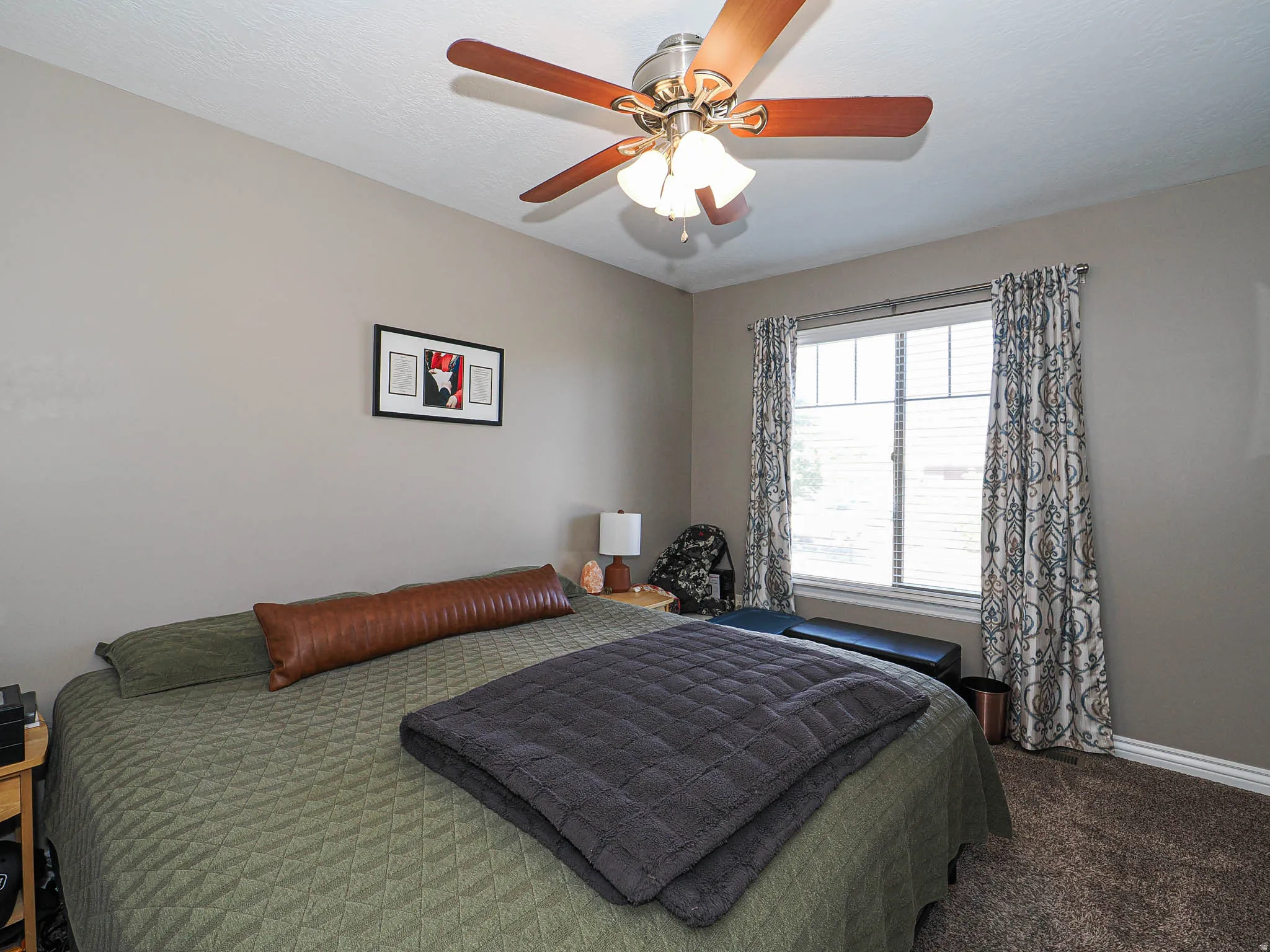 Bedroom featuring a ceiling fan with light fixture, window with blinds, neutral wall paint, and textured carpeting