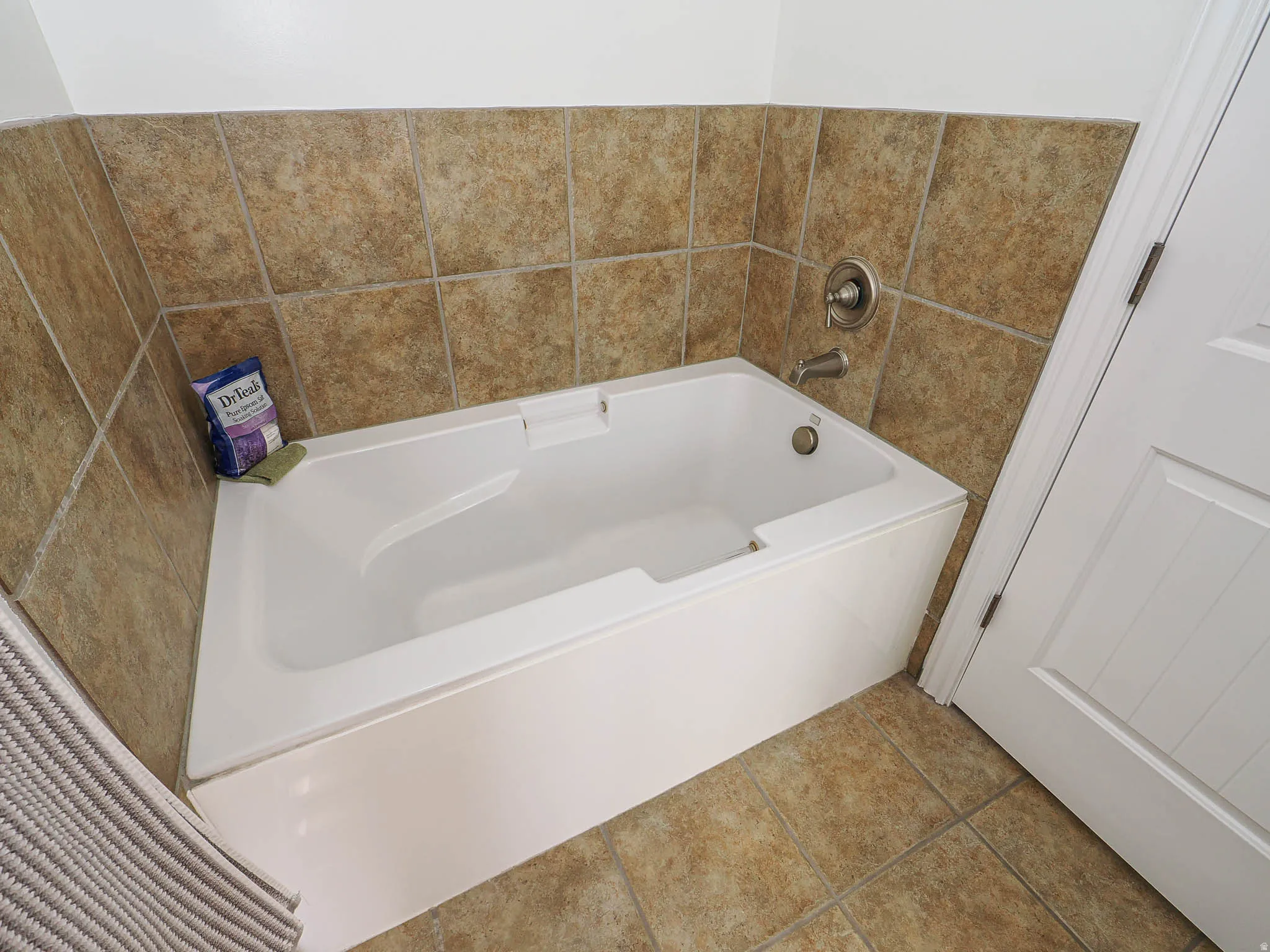 White soaking tub with brushed nickel fixtures, surrounded by neutral-toned tiled walls and flooring