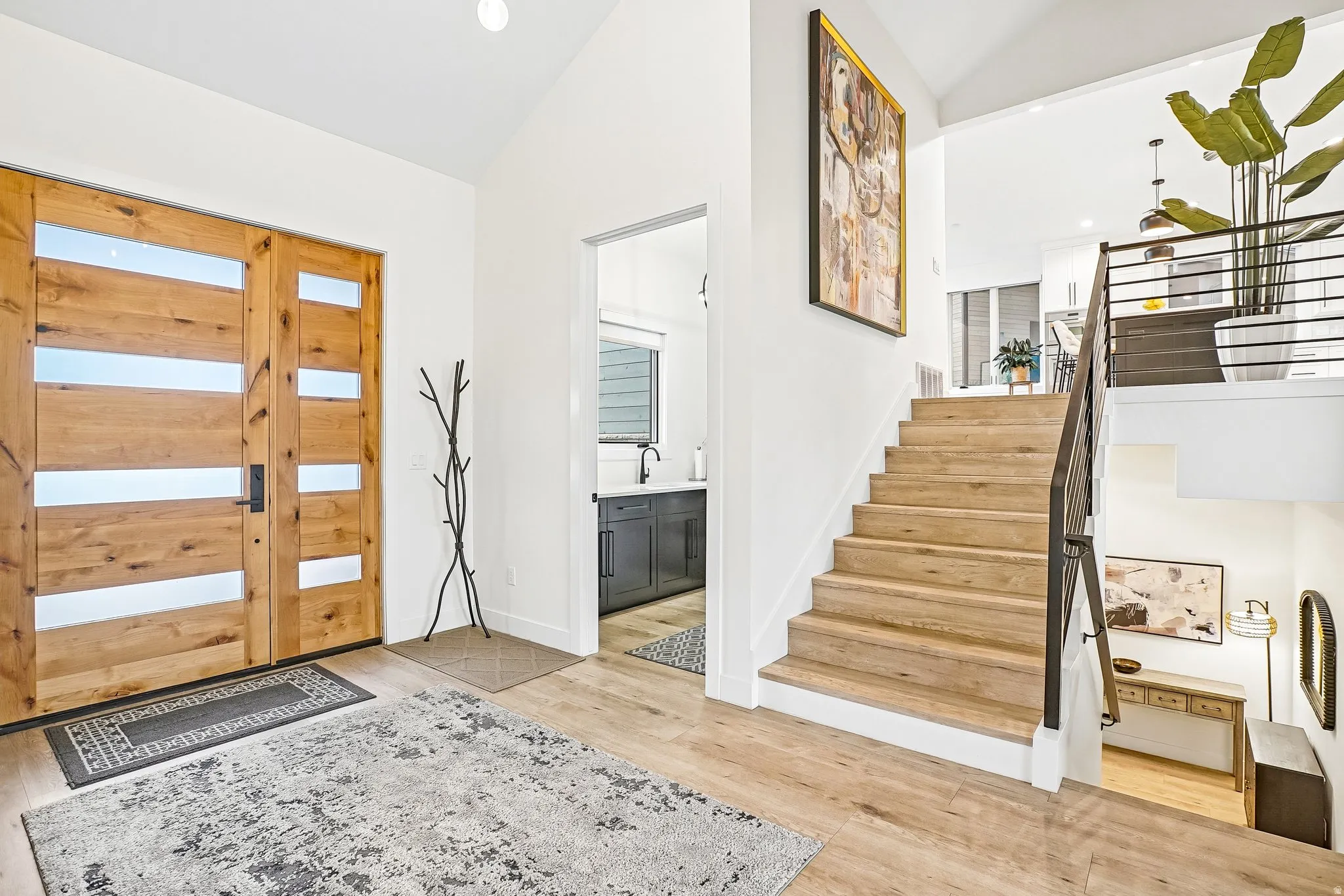 Double wood entry doors with horizontal frosted glass inserts open to a bright foyer featuring wood-finish flooring, a contemporary black metal staircase railing, and a vaulted ceiling