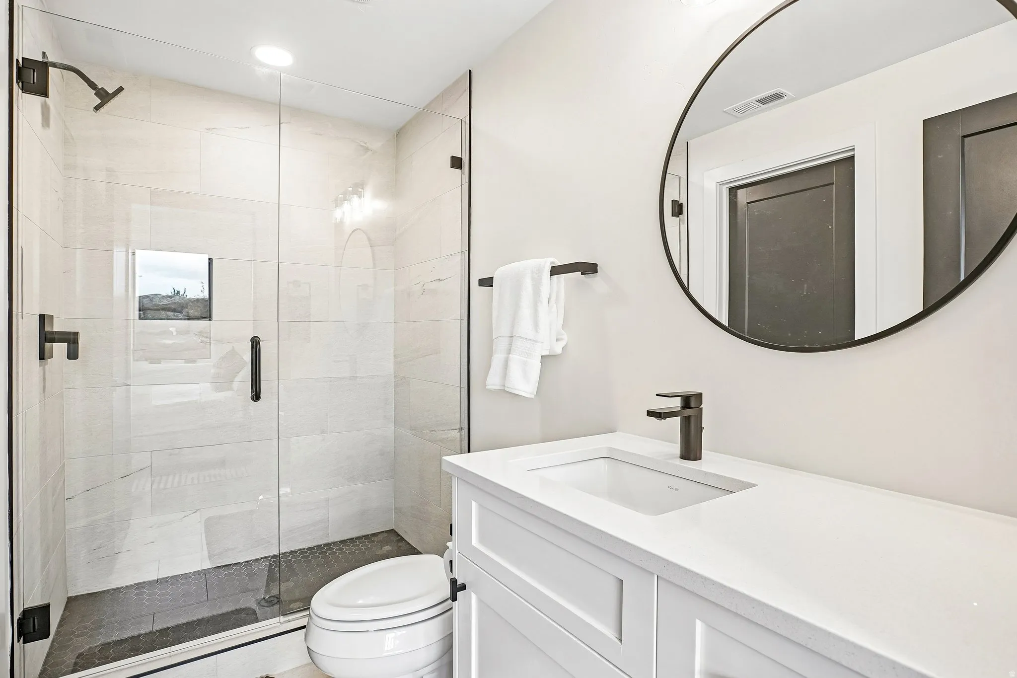 Modern bathroom featuring a frameless glass shower enclosure, white subway tile shower walls, dark tile shower pan, white vanity with integrated sink, and a round black-framed mirror