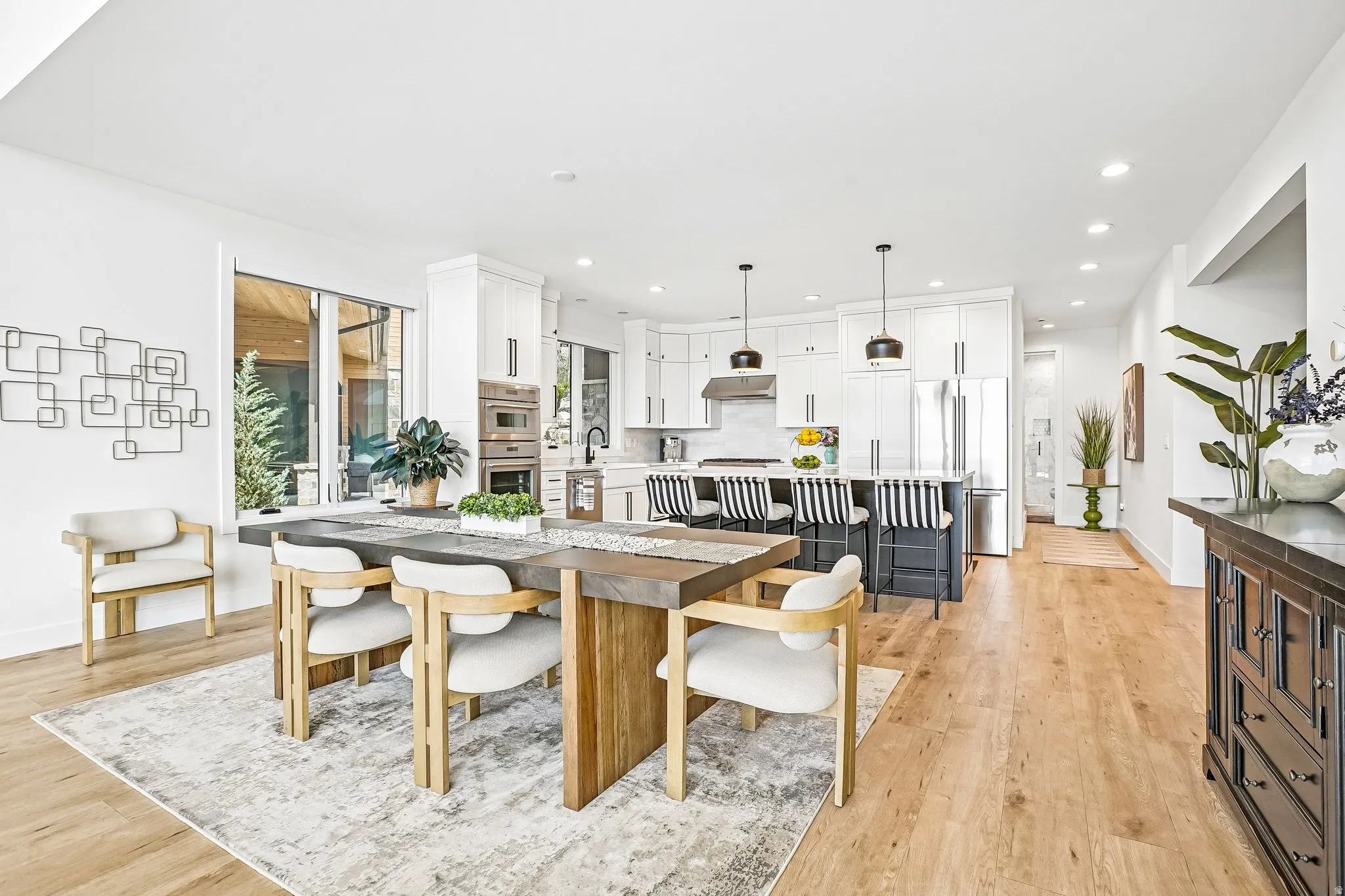 Open-concept kitchen and dining area featuring light wood-finish flooring, white cabinetry, stainless steel appliances, a center island with pendant lighting, and abundant natural light from large windows