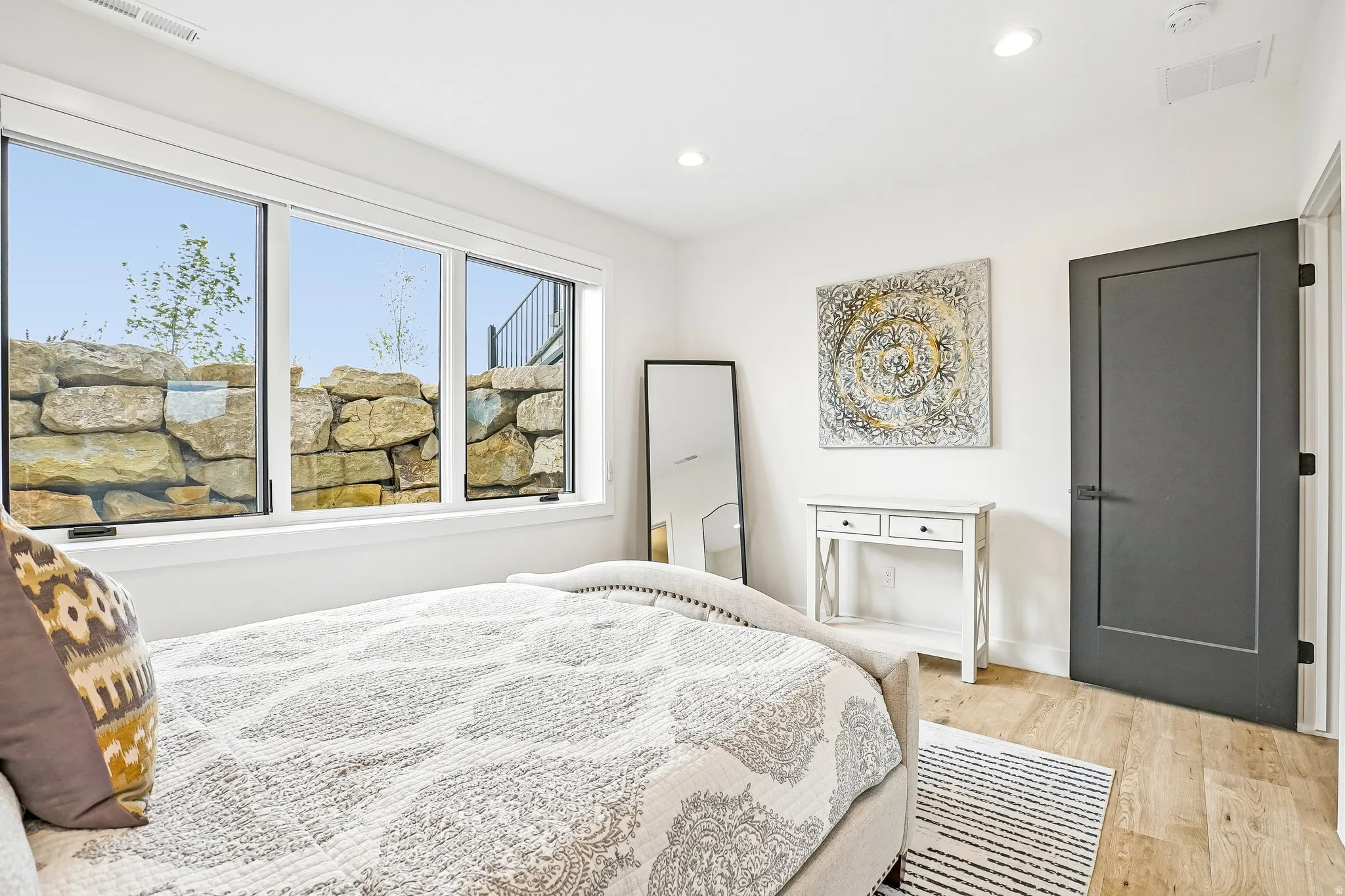 Large window overlooking a natural stone retaining wall, light wood-finish flooring, recessed lighting, and a dark paneled interior door