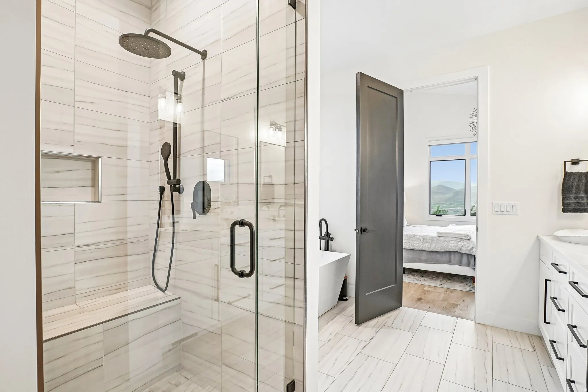 Spacious bathroom featuring a glass-enclosed shower with bench seating and a recessed niche, matte black fixtures, an adjacent freestanding soaking tub, and a vanity with a white countertop and dark hardware