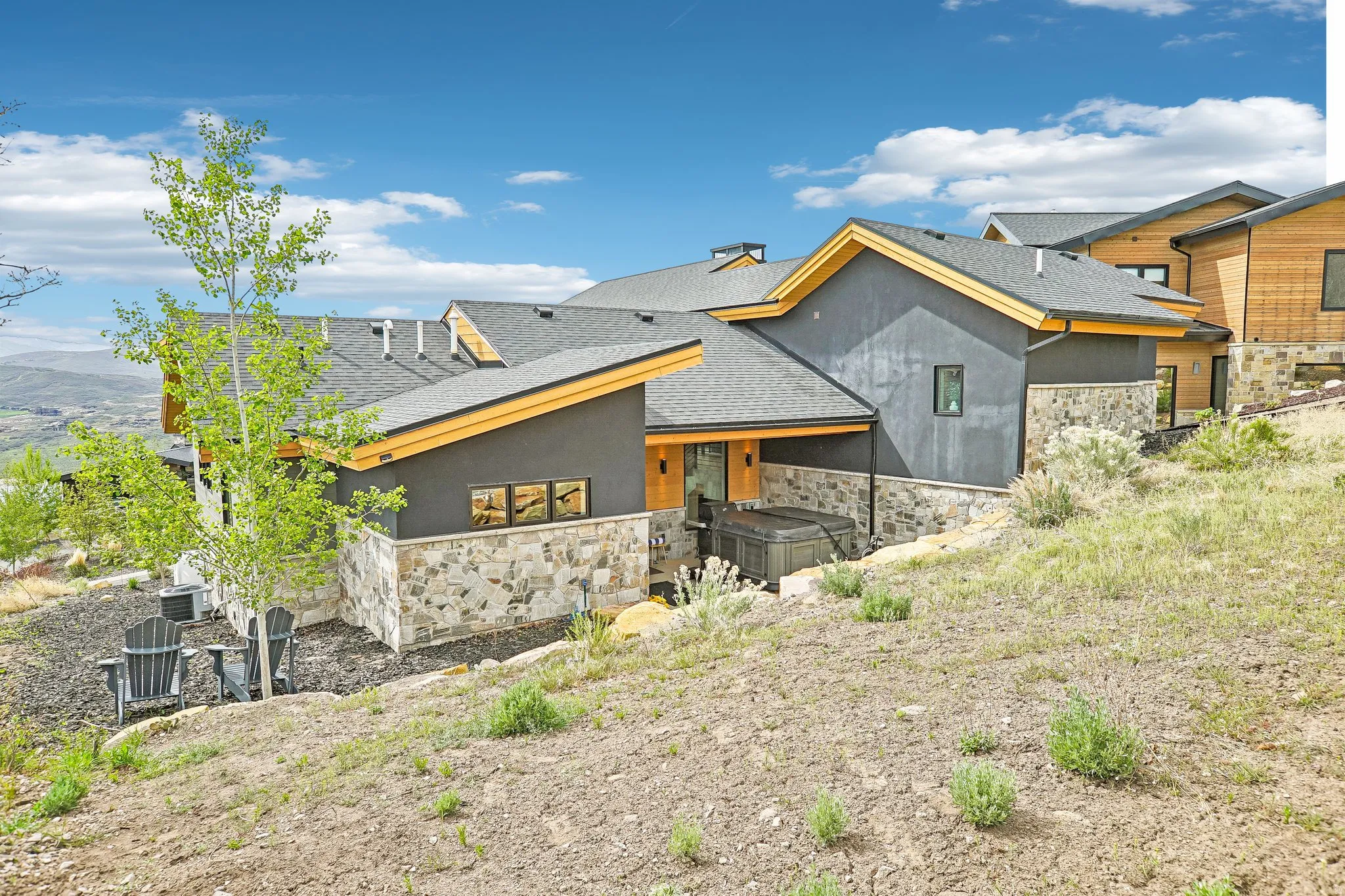 Modern exterior featuring dark gray stucco siding, natural wood accents, a stone foundation, a dark shingle roof, and a built-in hot tub