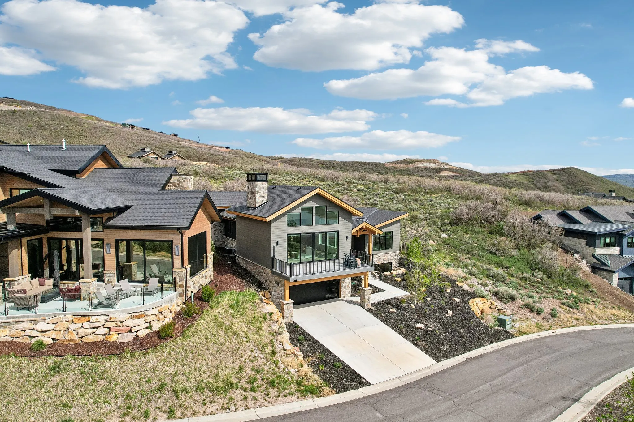 Modern mountain home featuring a stone and wood facade, a prominent stone chimney, an integrated garage, a spacious outdoor patio with a glass railing, and a sloped, natural landscape