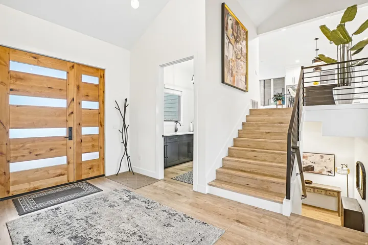 Double wood entry doors with horizontal frosted glass inserts open to a bright foyer featuring wood-finish flooring, a contemporary black metal staircase railing, and a vaulted ceiling