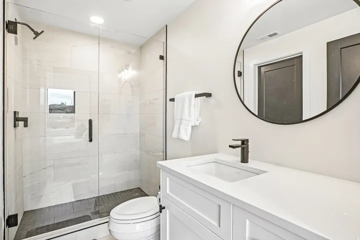Modern bathroom featuring a frameless glass shower enclosure, white subway tile shower walls, dark tile shower pan, white vanity with integrated sink, and a round black-framed mirror