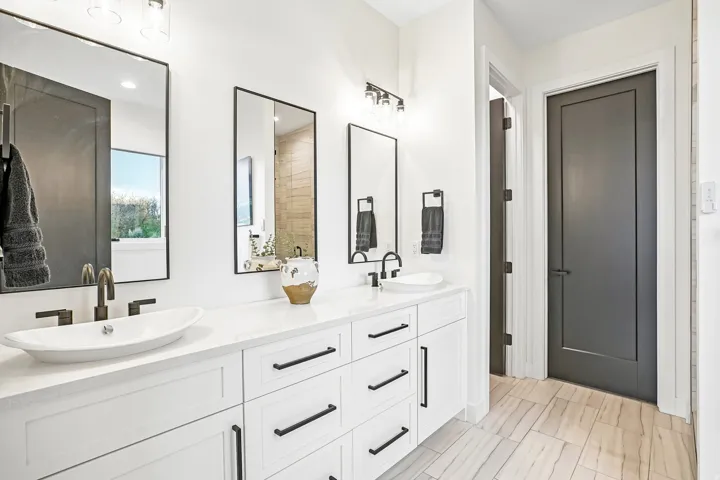 Dual vanity with white cabinetry and matte black hardware, white vessel sinks with wall-mounted bronze-finish faucets, framed mirrors, and wood-finish flooring
