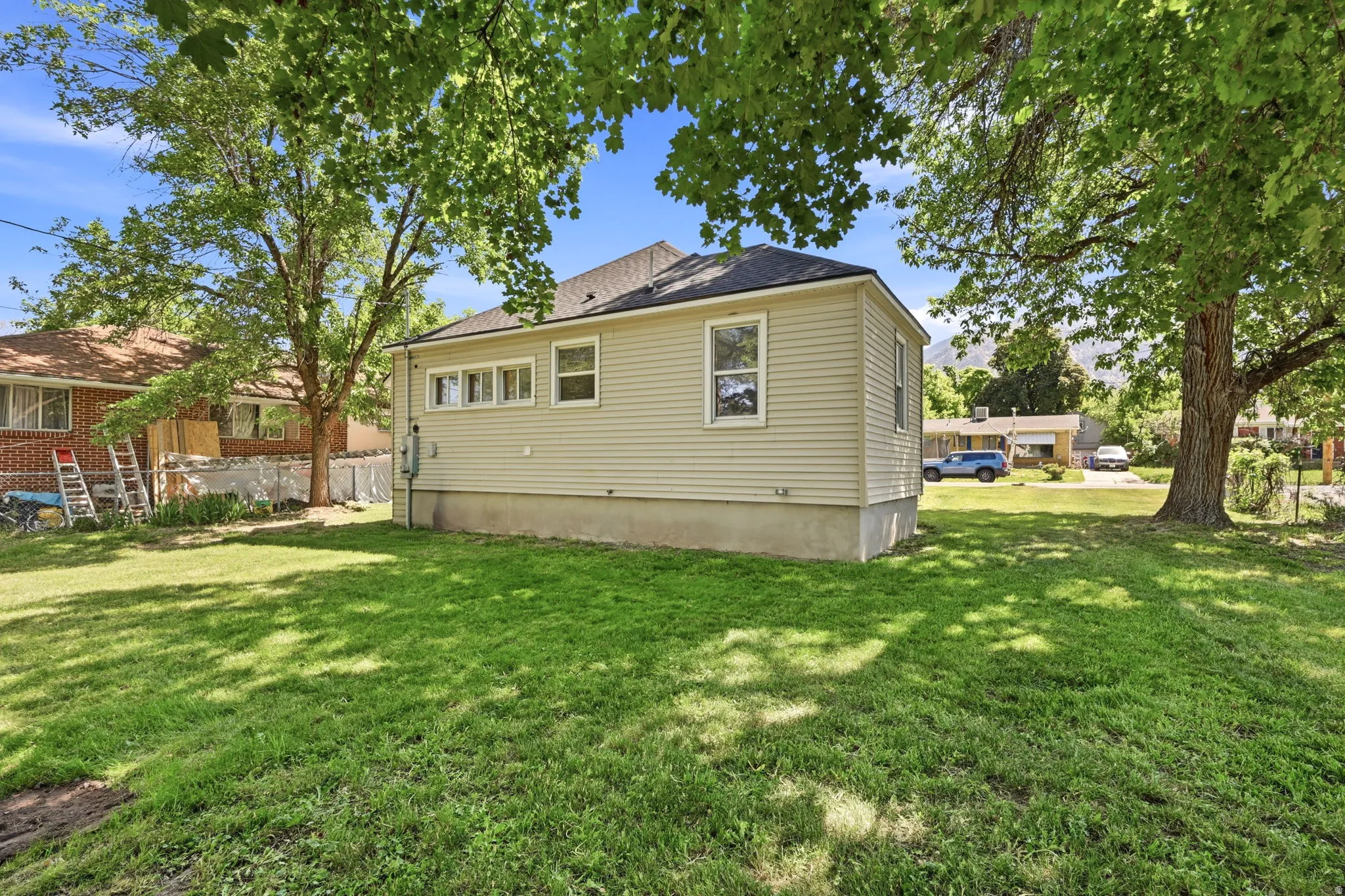 Expansive side yard featuring a manicured lawn, established shade trees, vinyl siding exterior, and a visible mountain backdrop