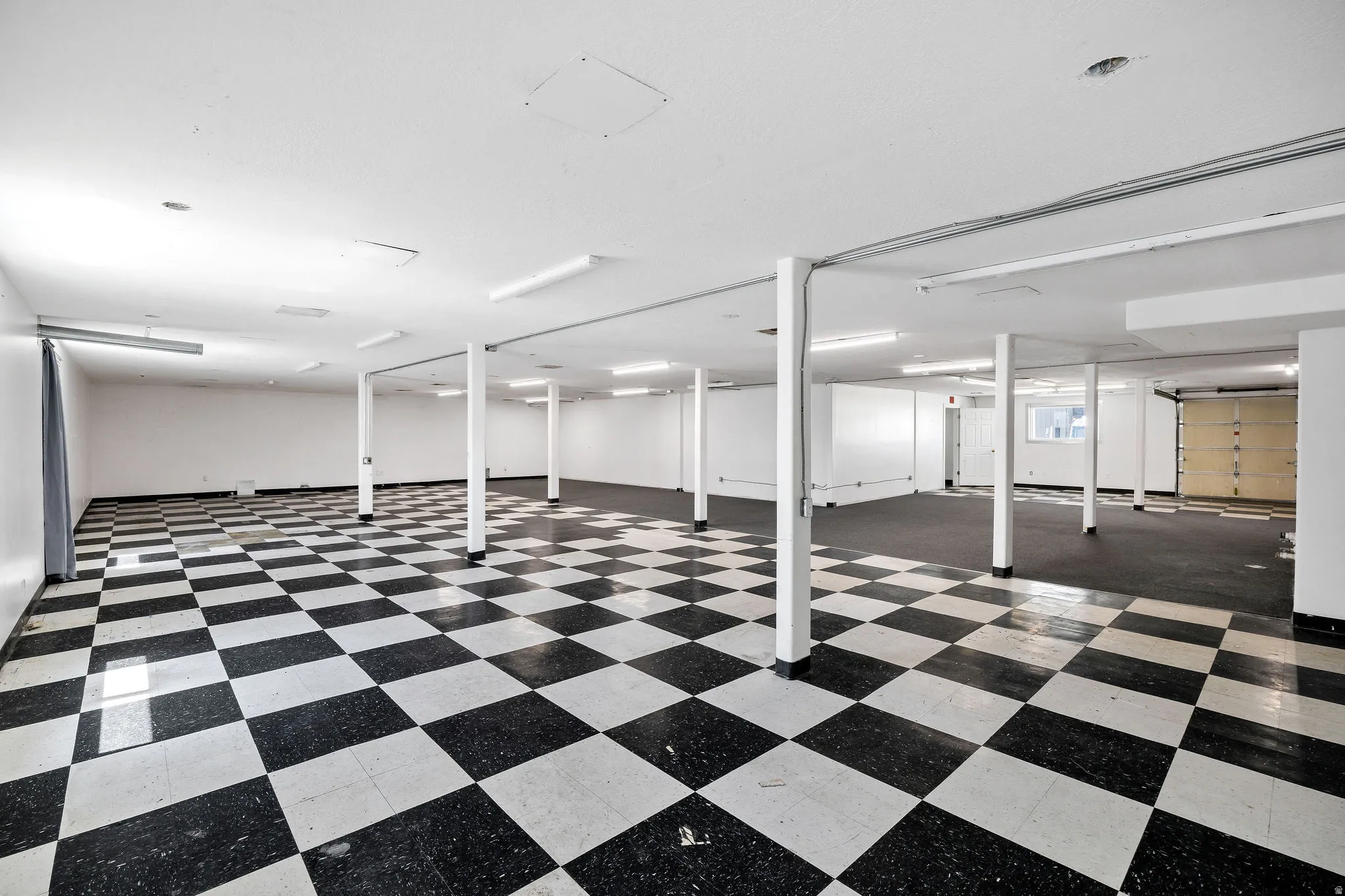 Expansive open space featuring a black and white checkerboard floor, bright overhead lighting, white support columns, and a large roll-up door