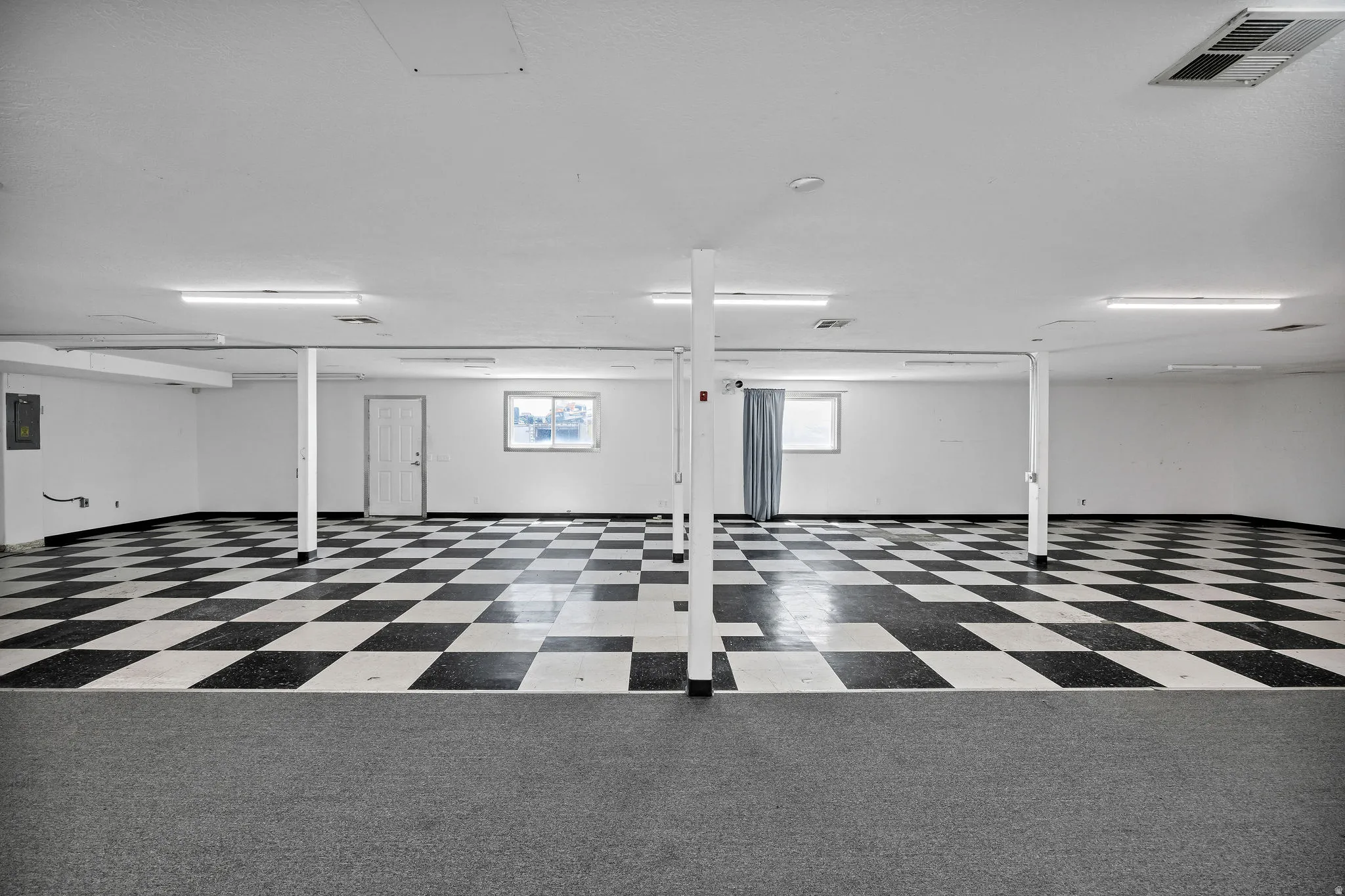 Spacious open area featuring black and white checkerboard flooring, white painted walls, and overhead fluorescent lighting