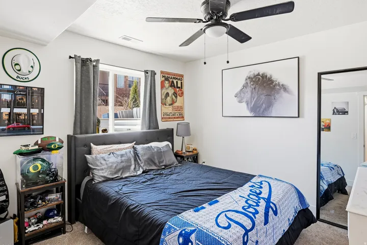 Carpeted bedroom featuring a window with dark gray curtains, a ceiling fan with integrated lighting, and white walls
