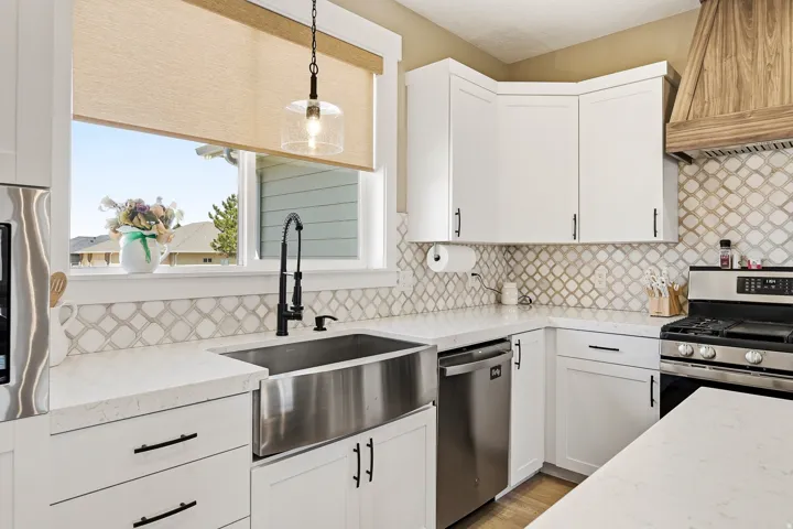 Bright kitchen featuring white cabinetry, light countertops, stainless steel appliances, a farmhouse sink with a gooseneck faucet, and a decorative tile backsplash