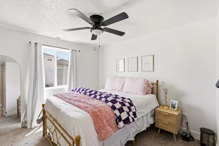 Bedroom featuring a window with white curtains, a ceiling fan with light fixture, and neutral-colored carpeting