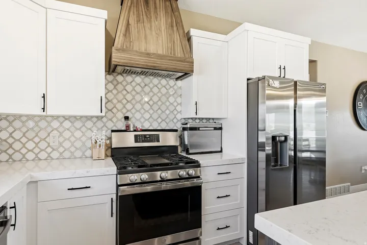 Stainless steel gas range with a wood-finish range hood, arabesque tile backsplash, white shaker cabinetry, and light-toned countertops
