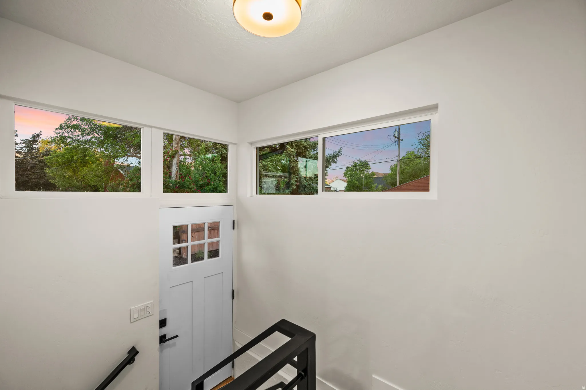 White painted door with upper window panes, complemented by multiple high-set horizontal windows, a flush-mount ceiling light fixture, and black metal handrailing to the finished basement