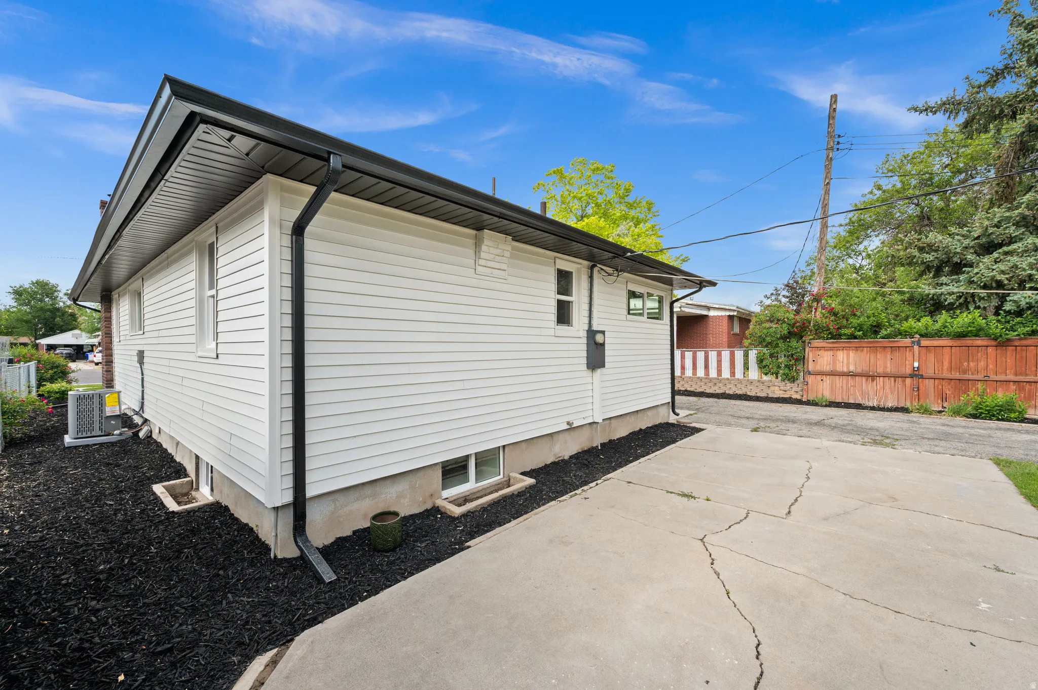 White siding exterior with black gutters and downspouts, featuring a concrete driveway, basement windows, and dark ground cover