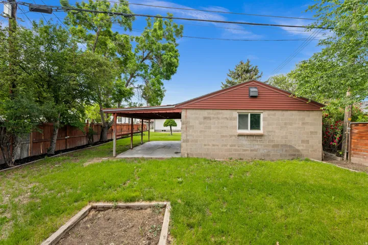 Expansive backyard featuring a concrete block structure with a red gable roof, an attached carport, and mature perimeter trees