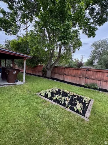 Expansive backyard featuring a lush lawn, a mature shade tree, a raised garden bed with planted greenery, and a wood fence enclosing the perimeter