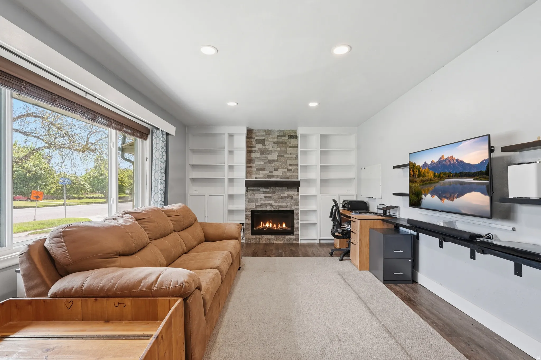 Living area featuring a stone-faced fireplace with a wood mantel, built-in shelving, recessed lighting, and large window with blinds