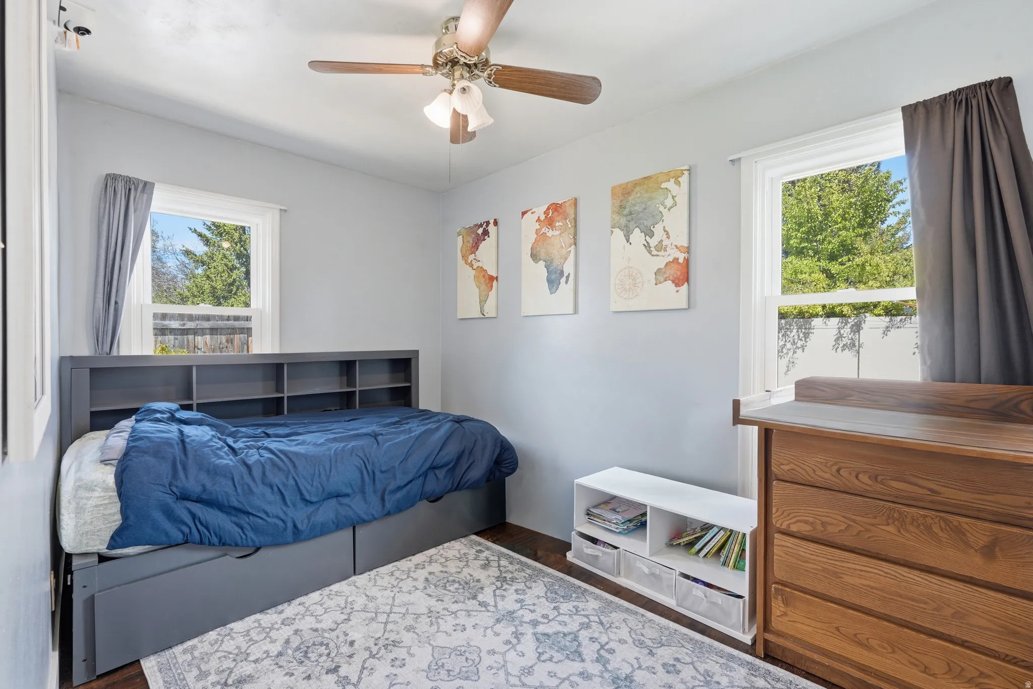 Bright bedroom featuring two large windows, a ceiling fan with integrated lighting, light gray wall paint, and wood-finish flooring