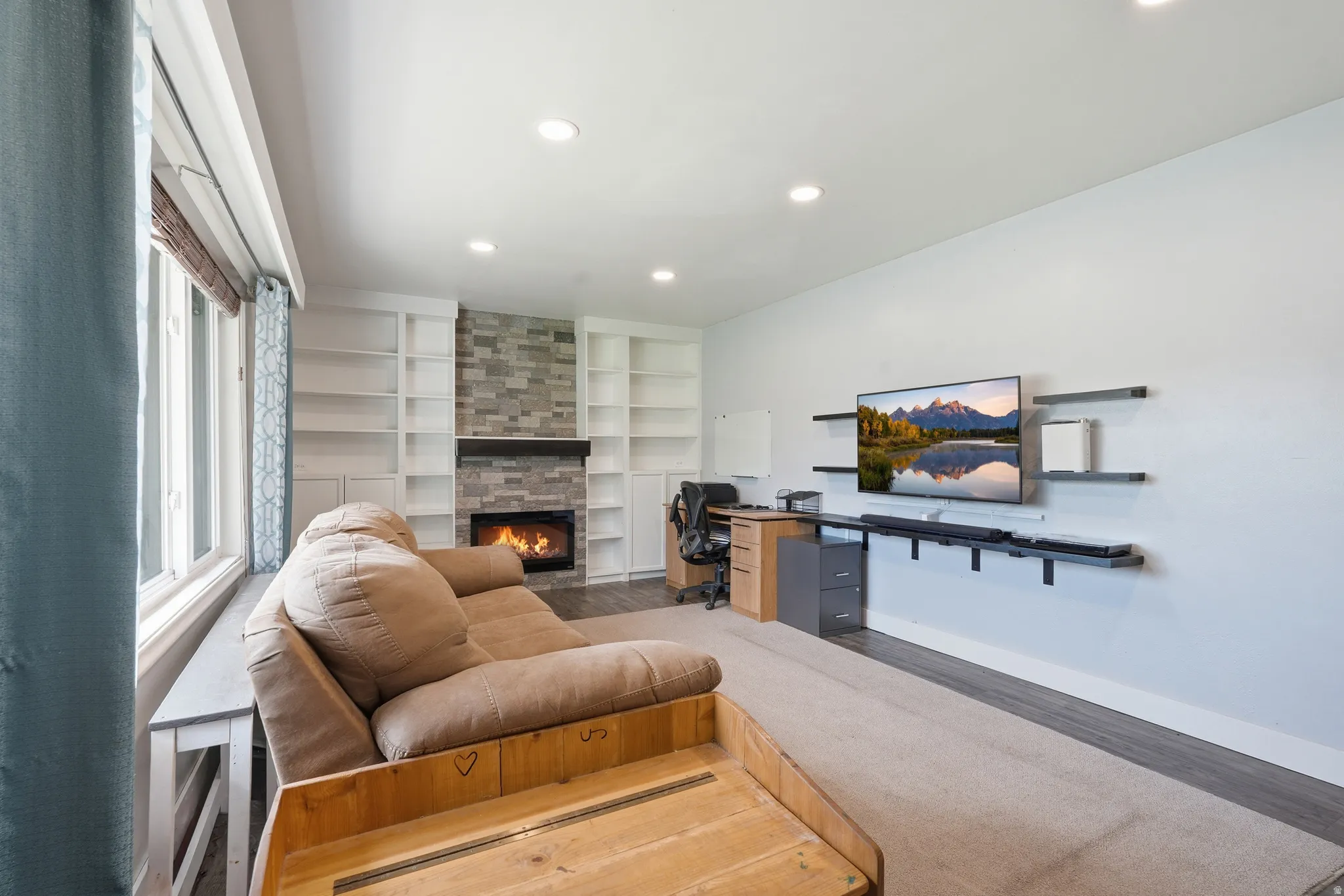 Spacious living area featuring a stone-clad fireplace with a dark wood mantel, built-in white shelving, recessed lighting, and large windows
