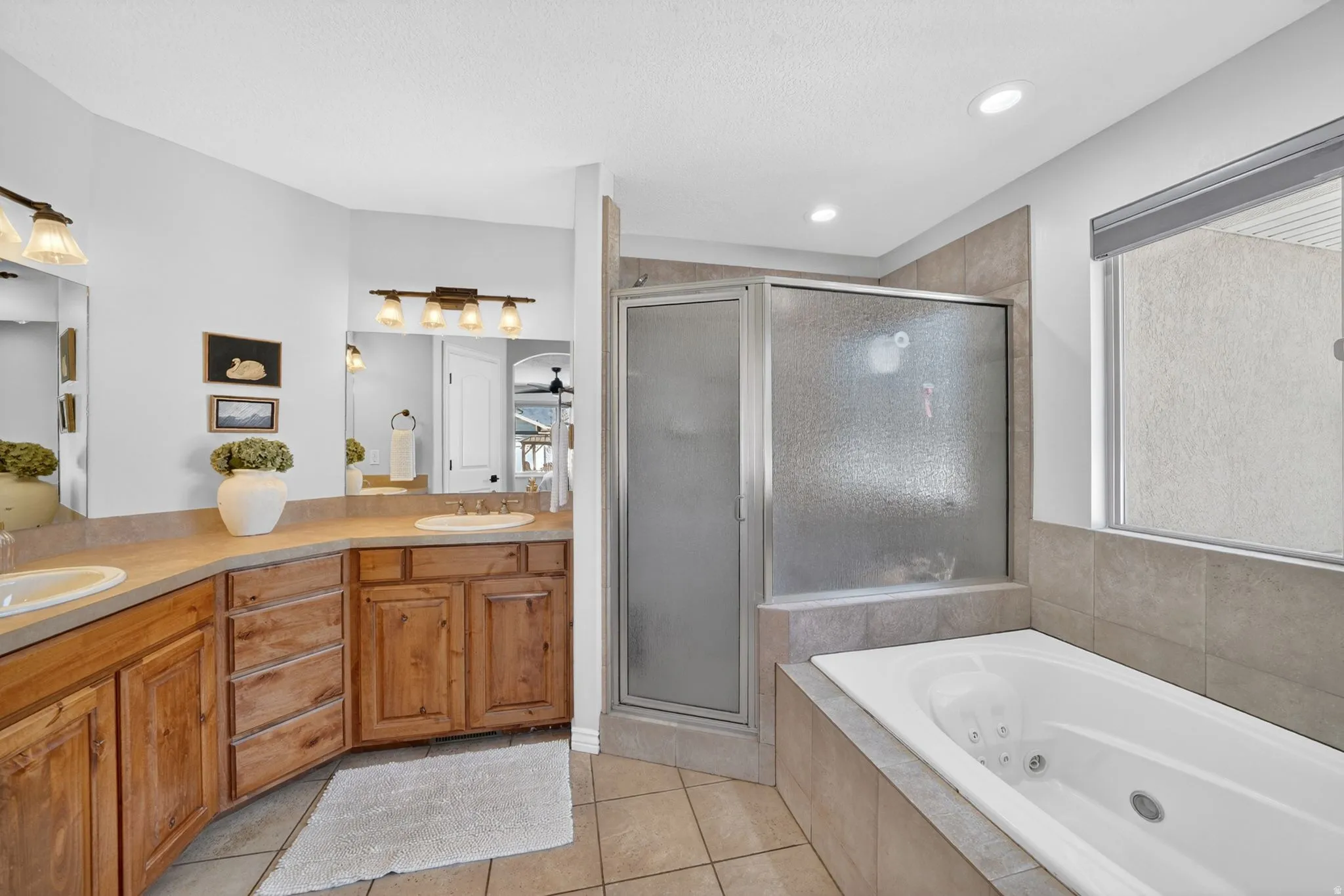 Bathroom featuring a jetted soaking tub, glass-enclosed shower, dual vanities with integrated sinks, wood-finish cabinetry, and neutral tile flooring
