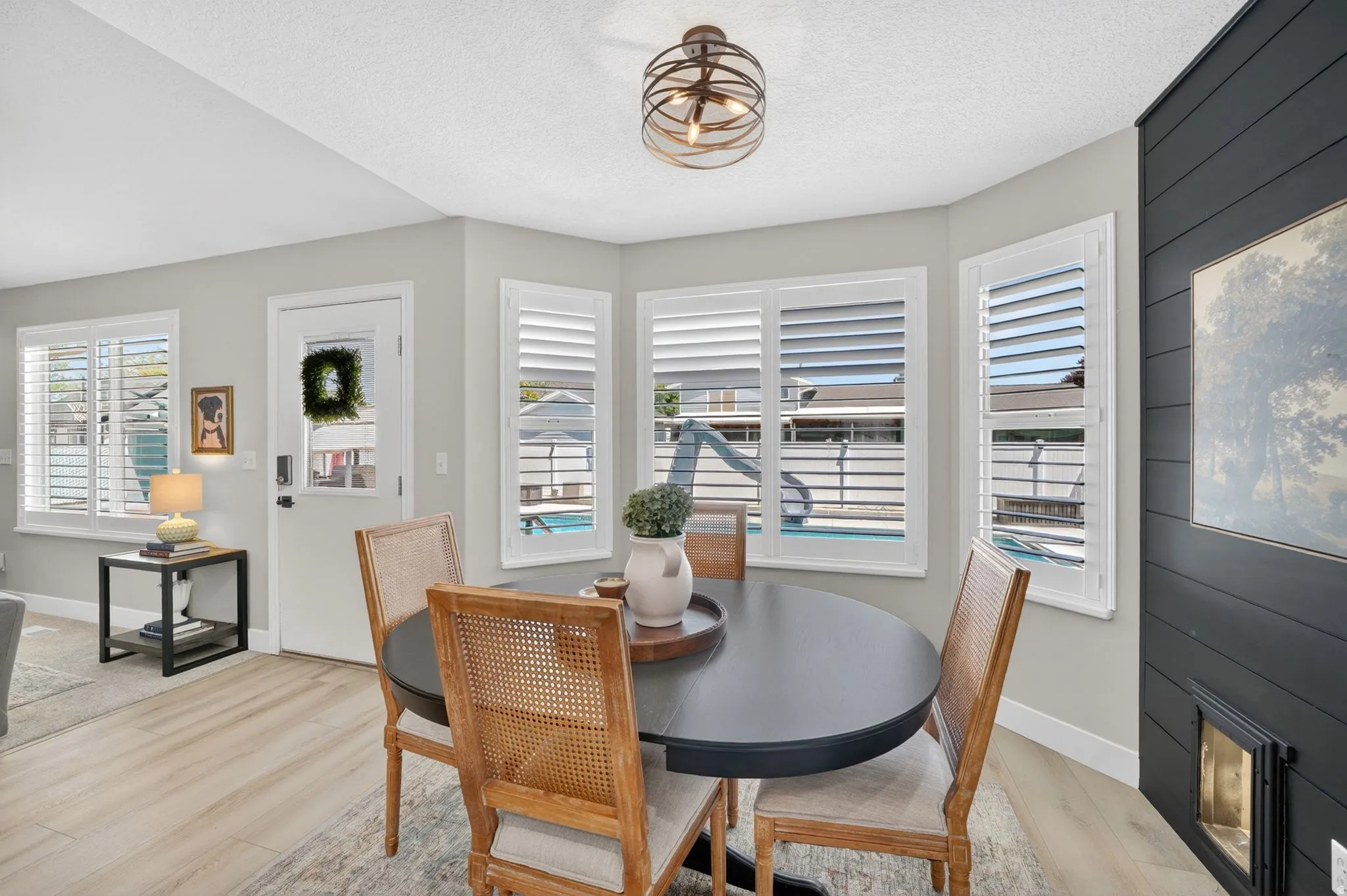 Dining area featuring a contemporary round table, light wood-finish flooring, a modern orb chandelier, multiple windows with white plantation shutters, and a dark accent wall with a built-in fireplace