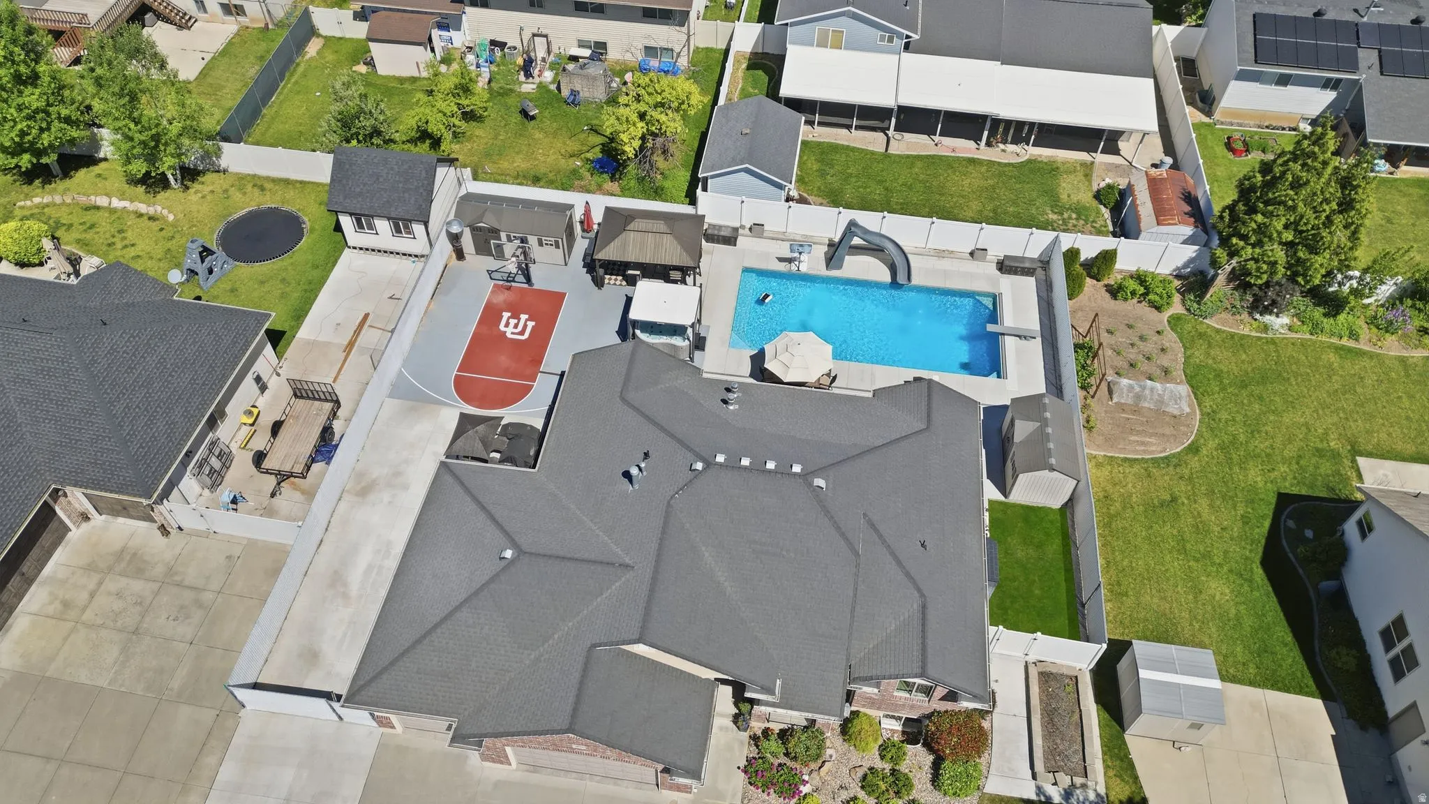 Aerial view showcasing a rectangular swimming pool with a slide, an enclosed sports court, a paved driveway, and multiple outbuildings