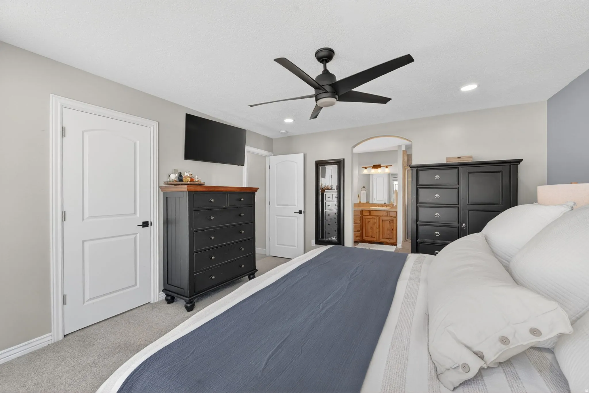 Carpeted bedroom featuring a contemporary ceiling fan with integrated lighting, recessed ceiling lights, an arched doorway, and a bathroom entry with a wood-finish vanity and mirror