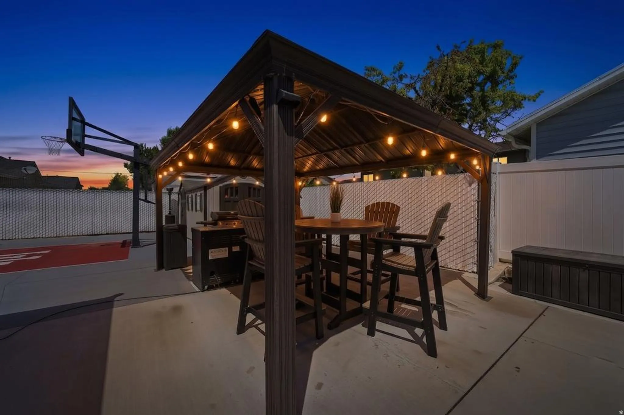 Outdoor gazebo with string lights, concrete patio, a basketball hoop with a painted court, and privacy fencing