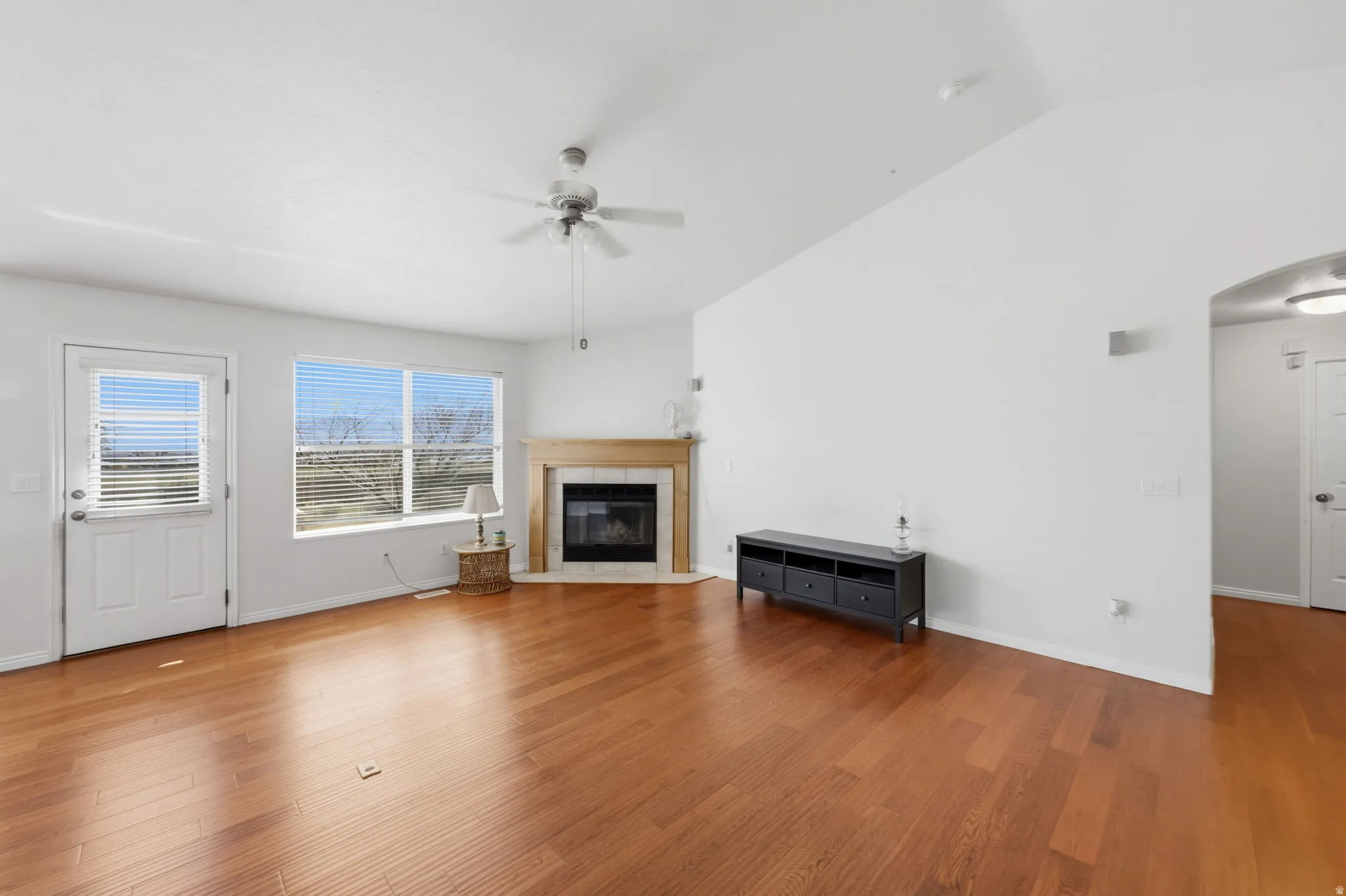 Living space featuring wood-finish flooring, a corner fireplace with a wood mantle, a ceiling fan, and large windows with horizontal blinds
