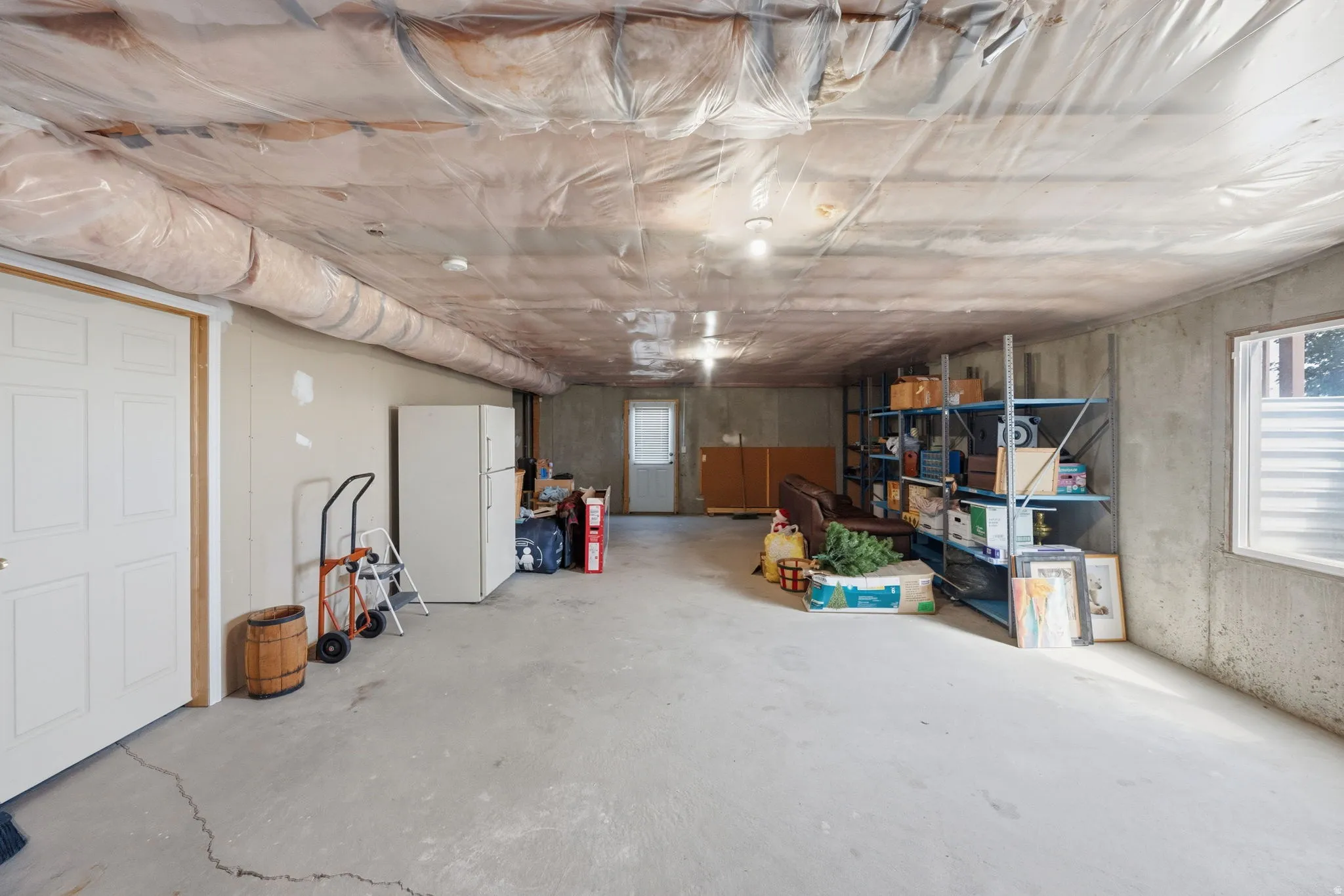 Expansive lower level featuring a concrete floor, exposed ceiling joists with insulation, two panel doors, a window with horizontal blinds, and concrete foundation walls